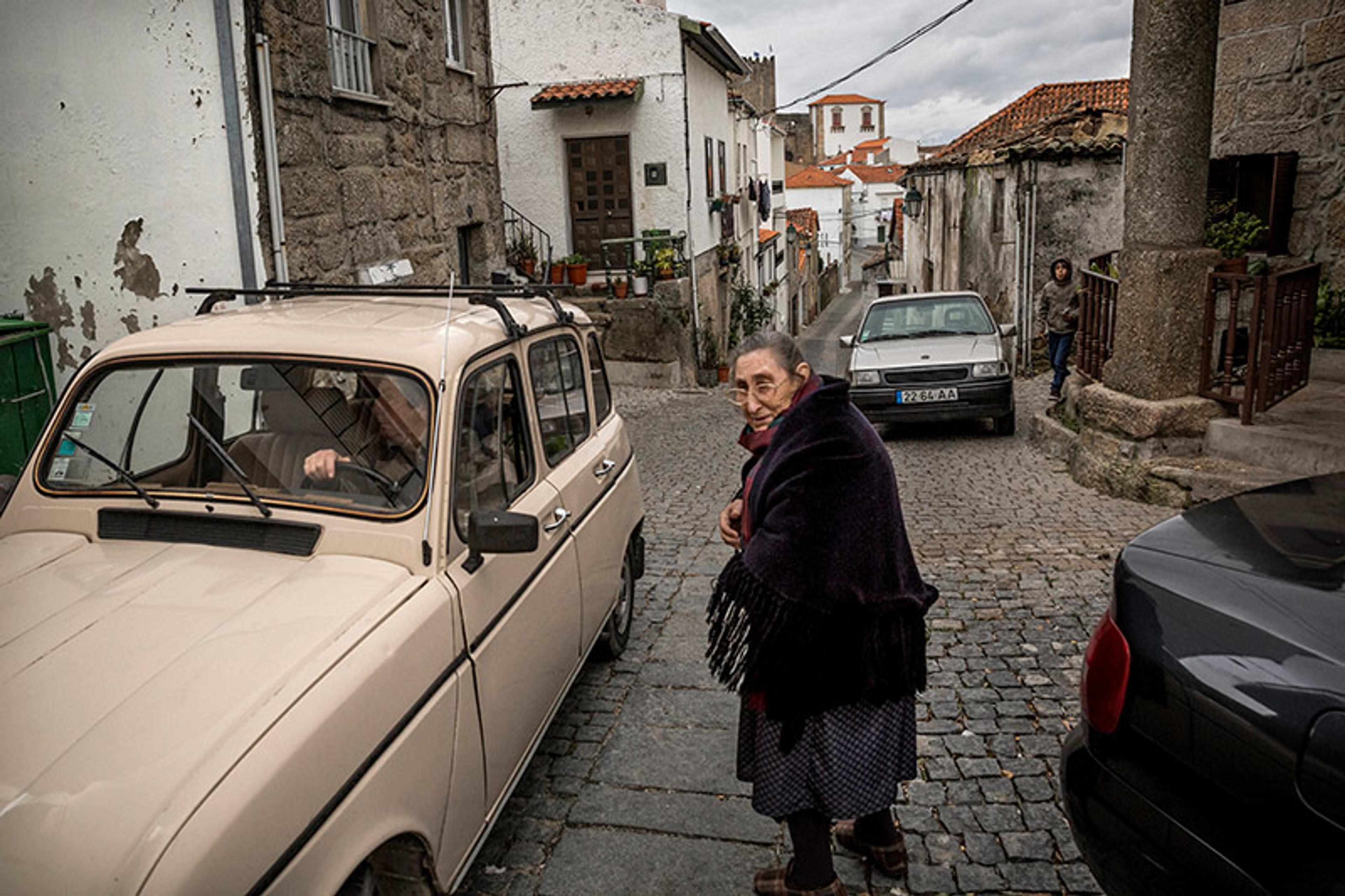 An elderly woman in a shawl on a cobblestone street, with parked cars and stone buildings in a village setting.