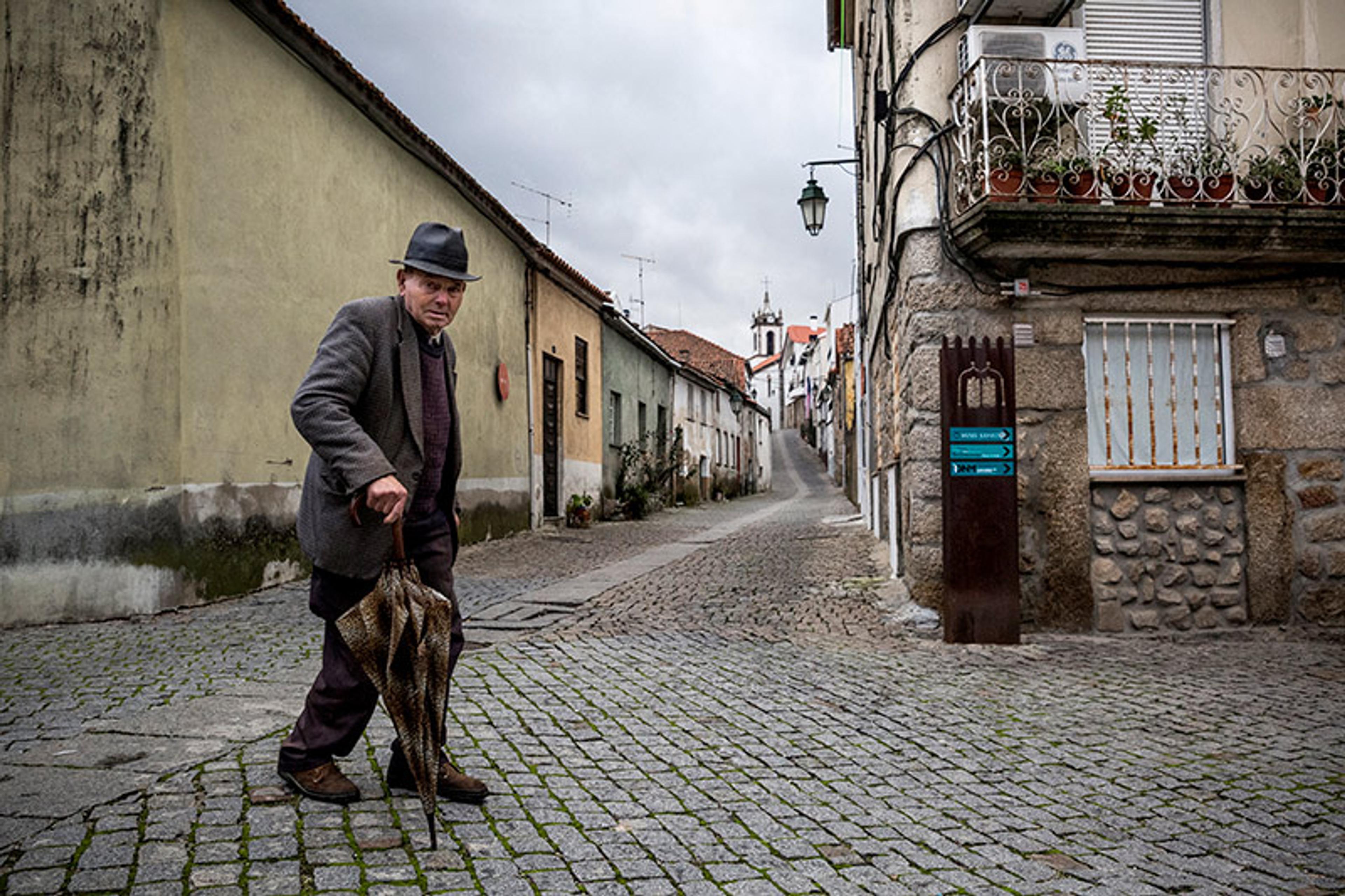 An elderly man walking on a cobblestone street in an old town, holding a folded umbrella with buildings and a church in the background.