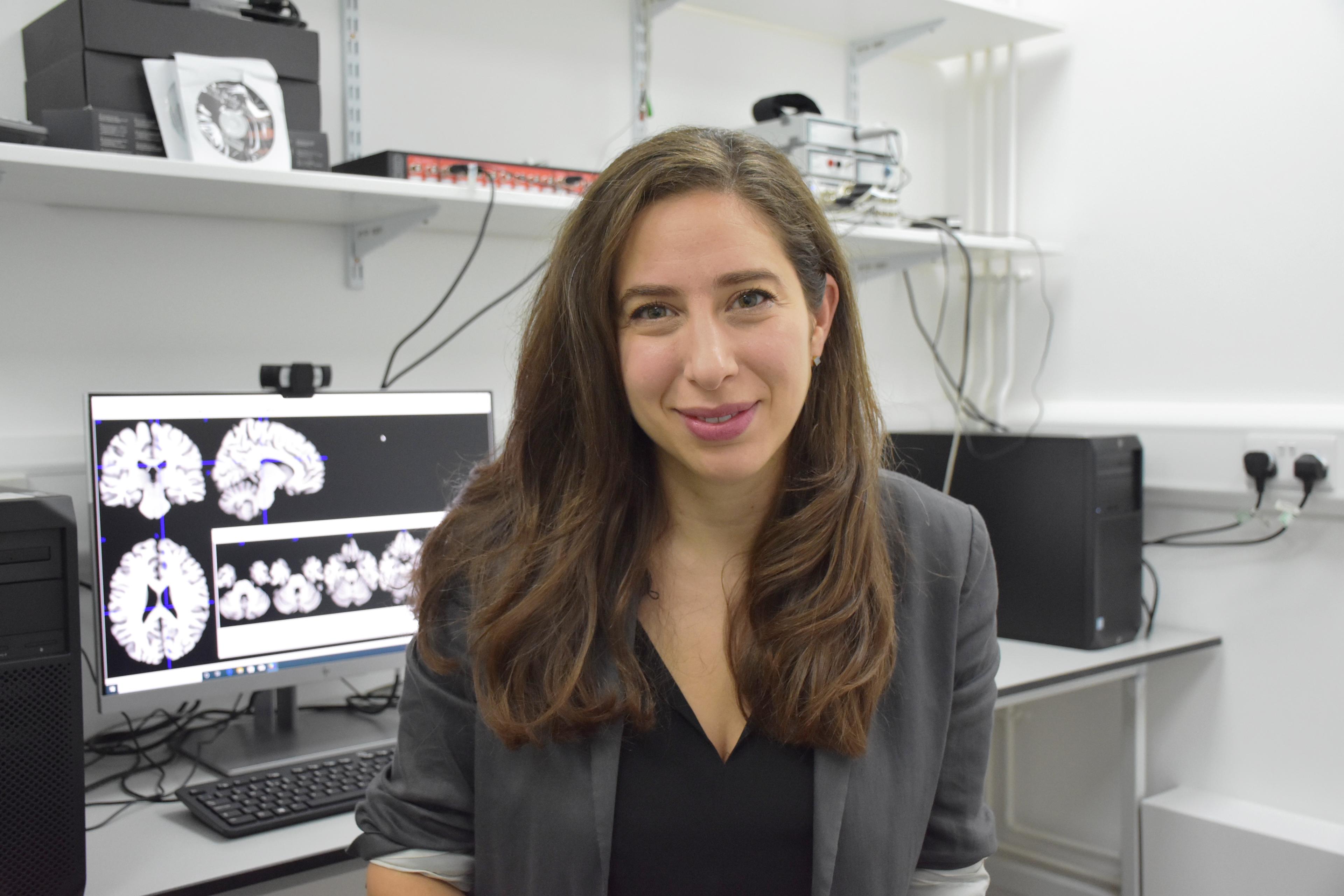 A woman with long hair in an office, with neurology brain scans displayed on a computer monitor behind her.