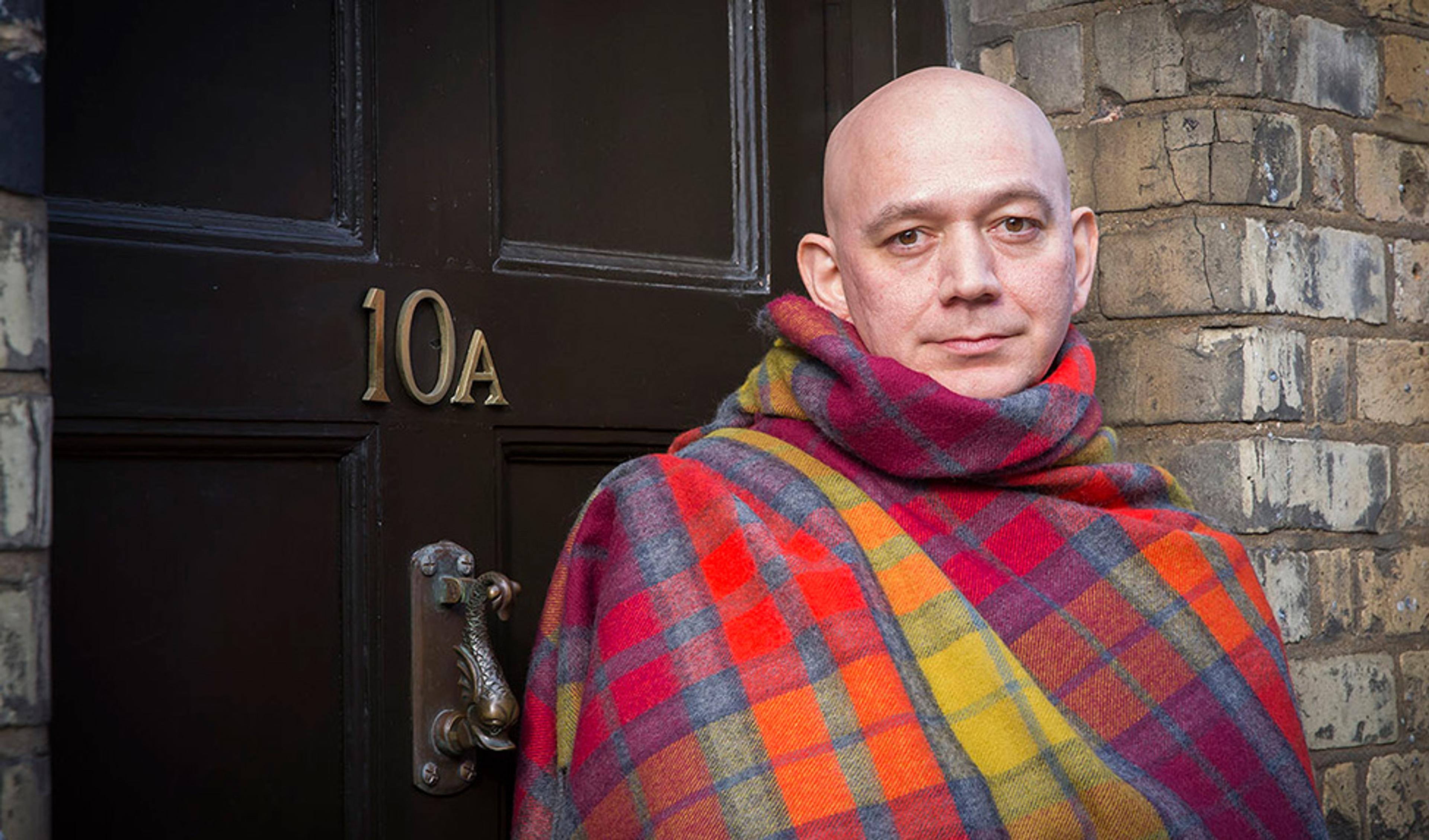 A bald man wrapped in a colourful plaid blanket standing by a door marked with 10A in gold, surrounded by a brick wall.