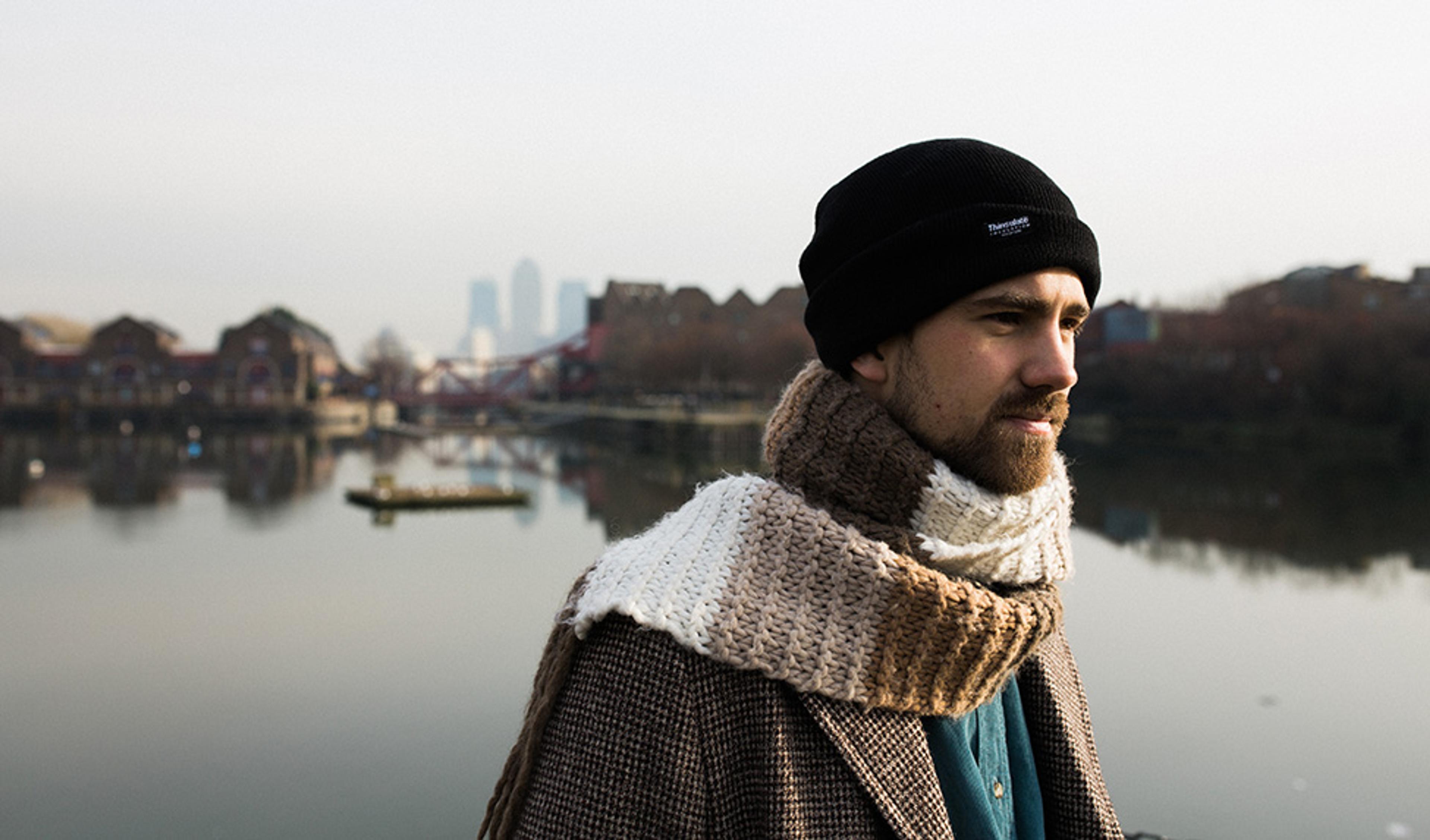 A man wearing a beanie and scarf standing by a calm river with buildings and a bridge in the background on a foggy day.