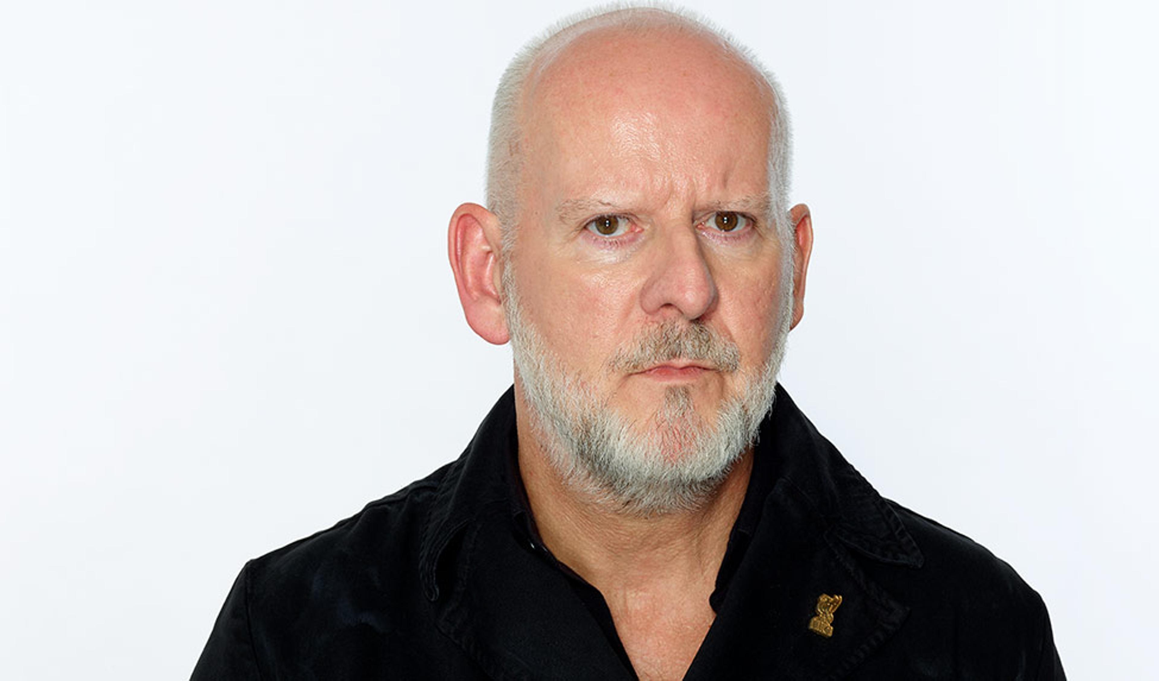 A bald man with a grey beard wearing a black jacket and a serious expression against a plain white background.