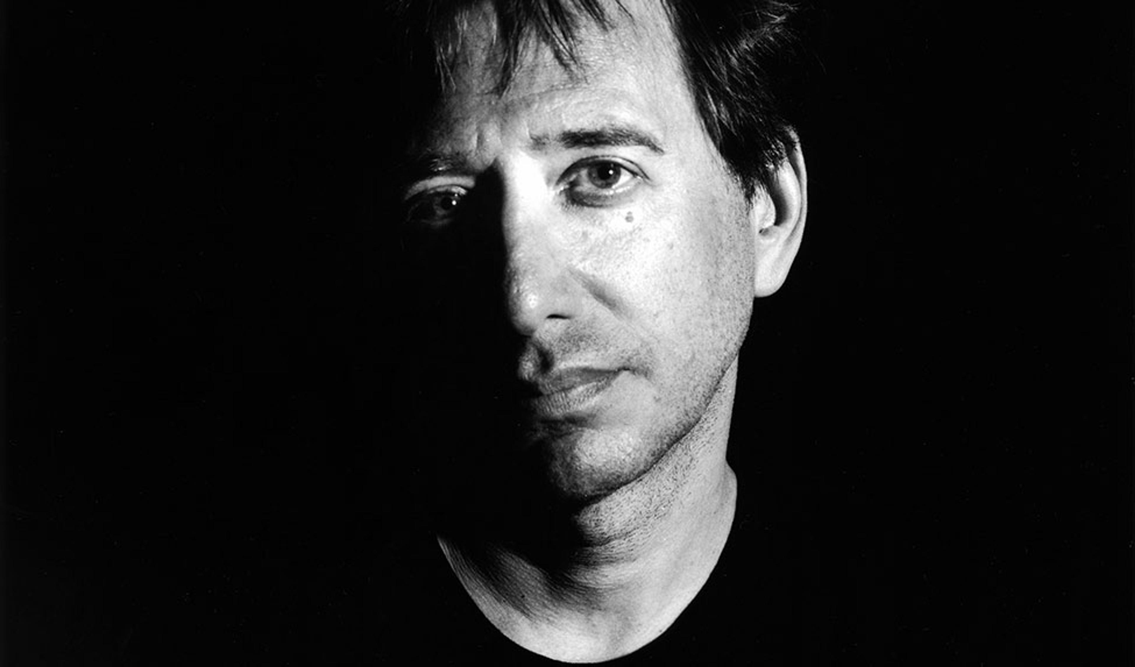 Black-and-white photo of a man with short hair in shadow, half of his face is brightly lit, set against a dark background.