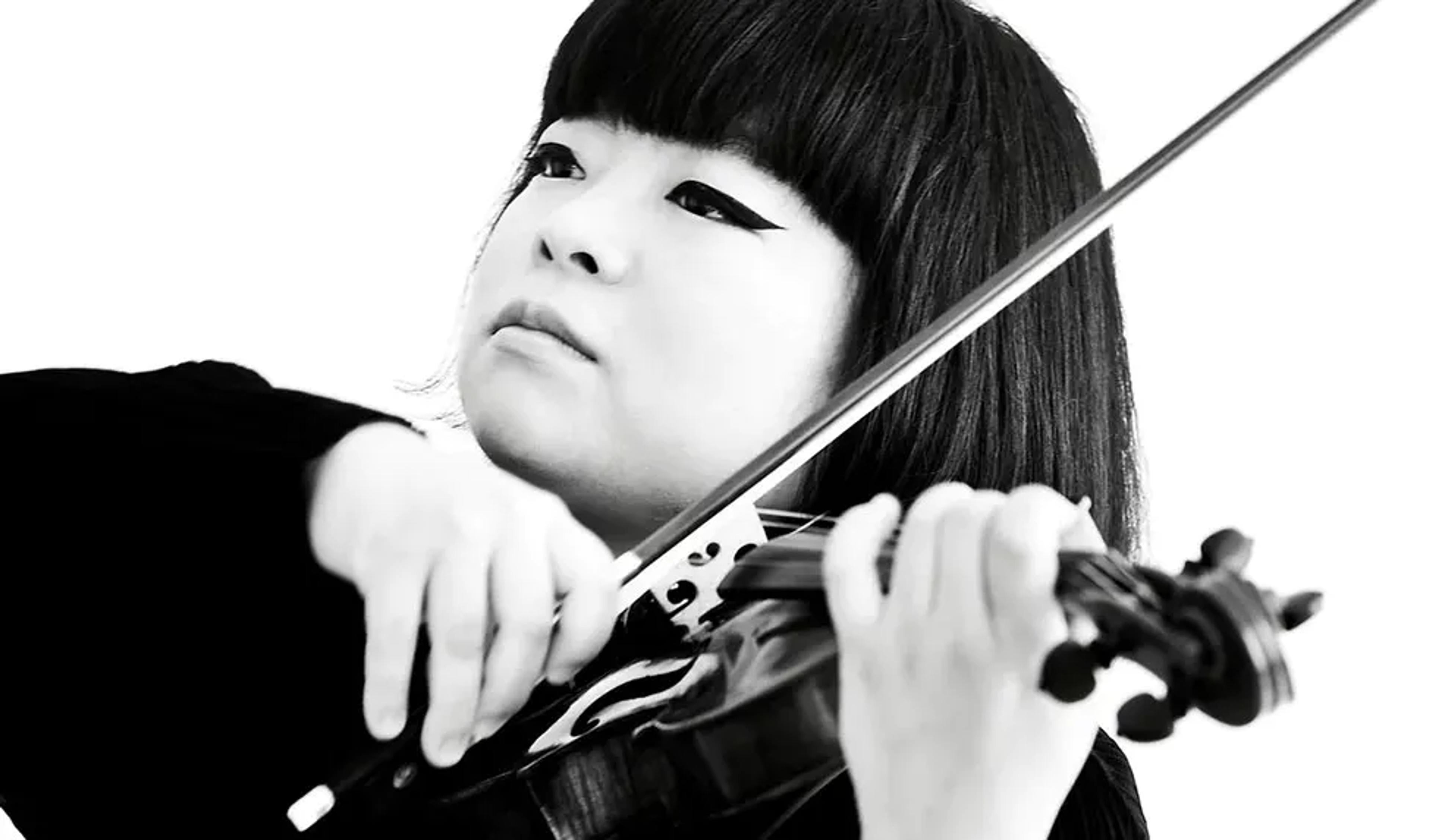 Black and white photo of a woman with short hair playing the violin, wearing dark eyeliner against a bright background.