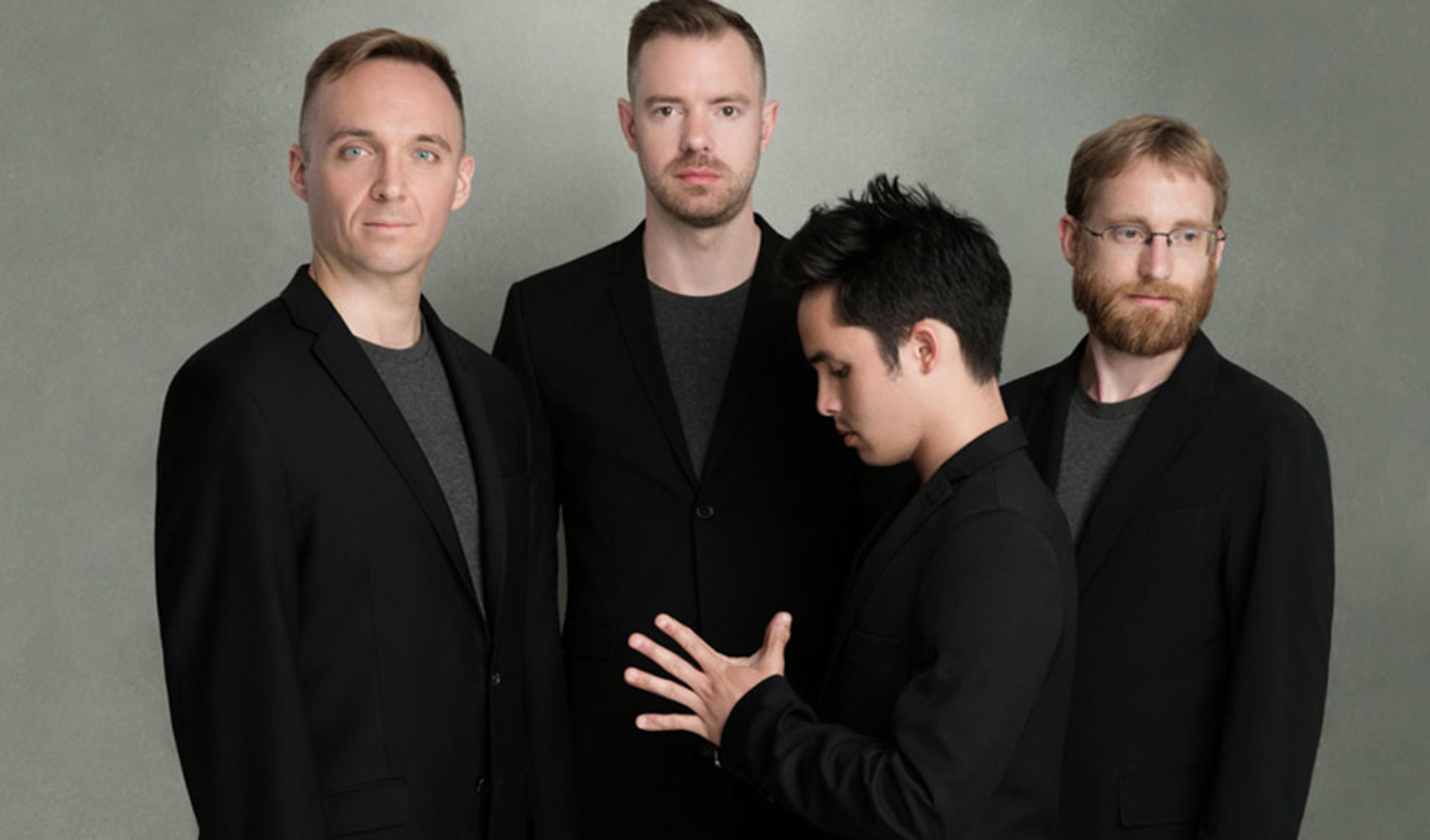 Four men in black jackets against a grey background. One man faces the camera, the second looks down, and two look ahead.