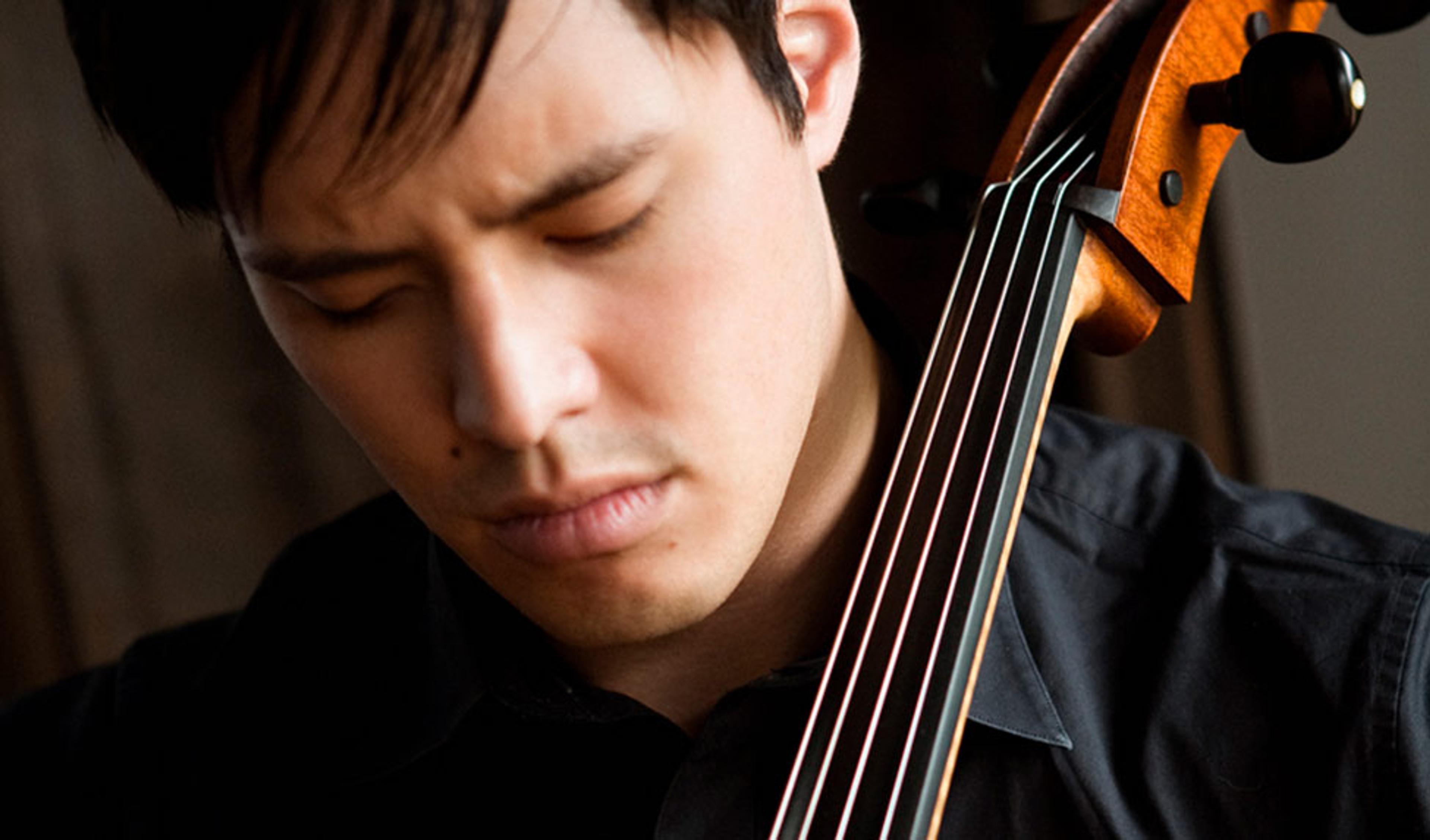 A young man concentrating while playing the cello, with the cello’s neck and strings visible in the foreground.