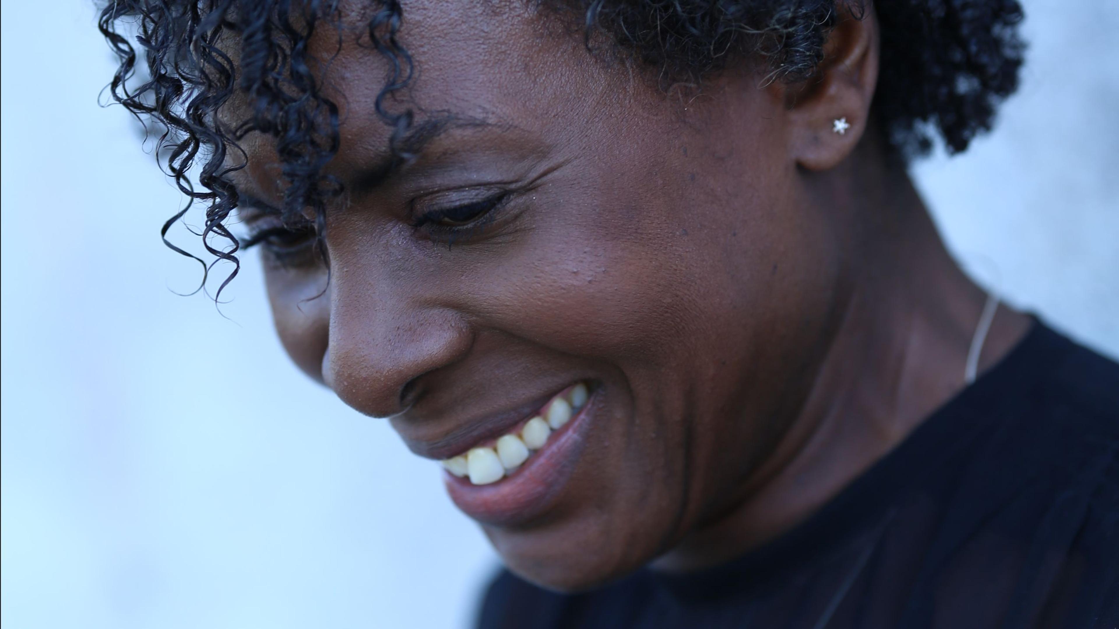 Close-up photo of a woman smiling, looking down with curly hair, wearing a star-shaped earring and a black top against a light background.