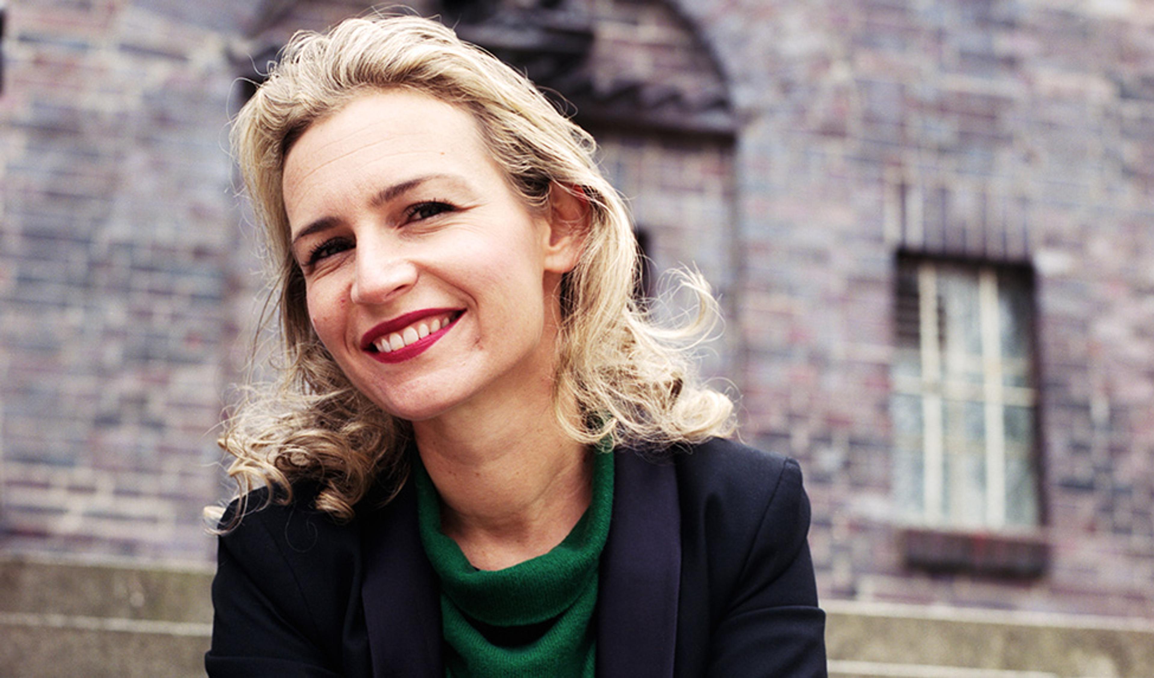 A smiling woman with blonde hair in a green top and black blazer, sitting in front of a blurred brick building.