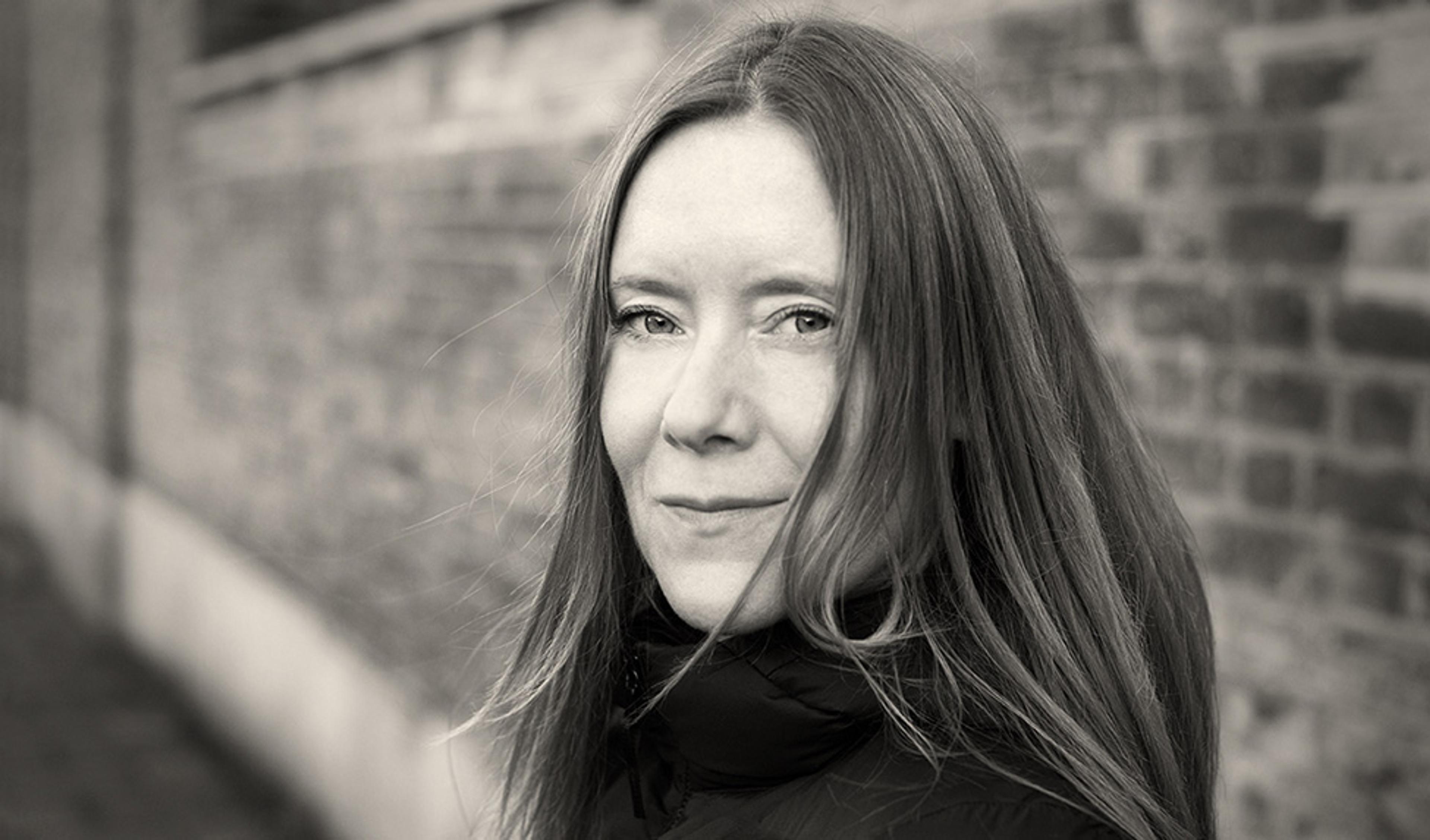 Black and white photo of a woman with long hair and a coat standing in front of a brick wall, looking into the camera.