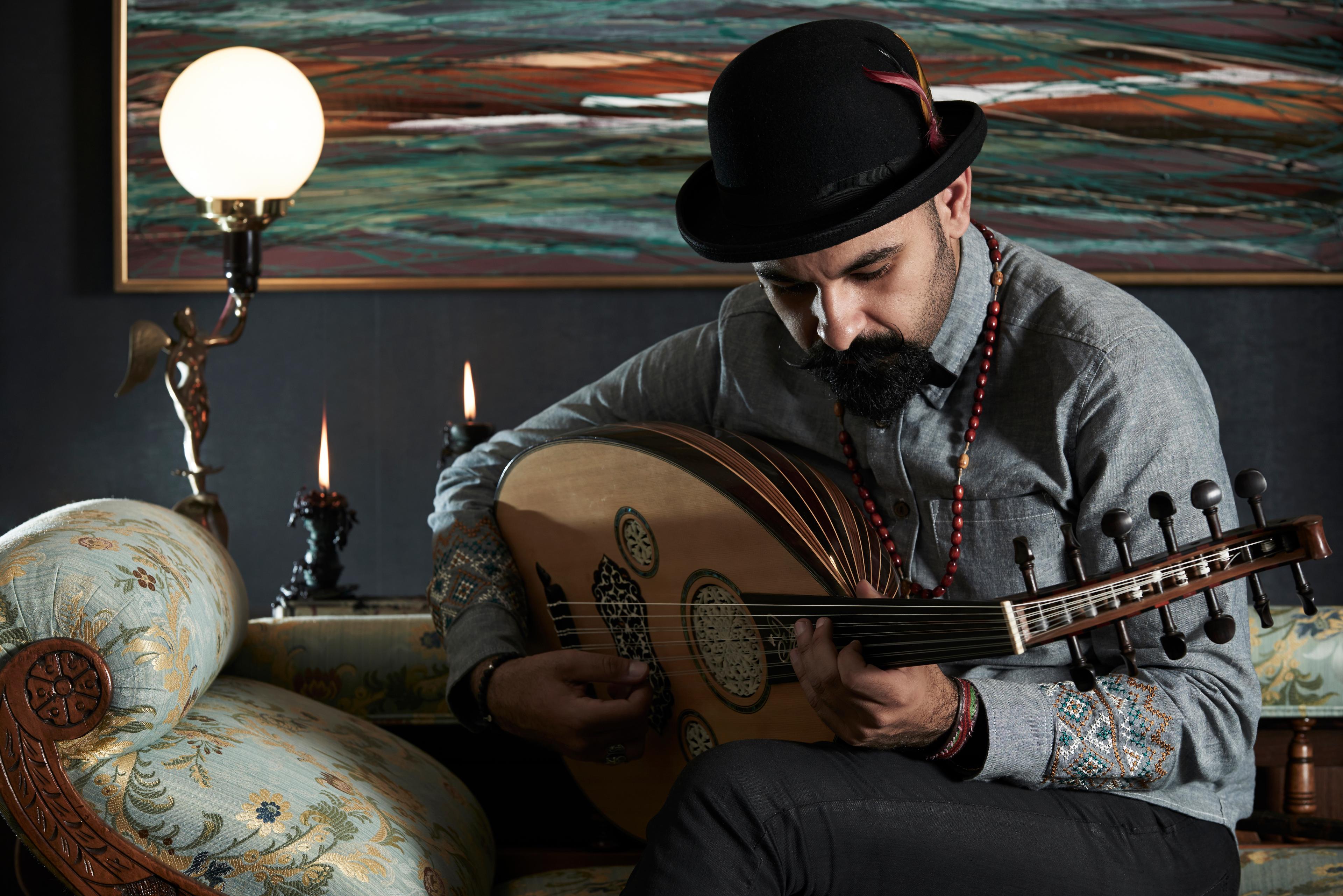 A man playing an oud with a modern painting, lamp and two candles in the background. He wears a hat and beaded necklace.