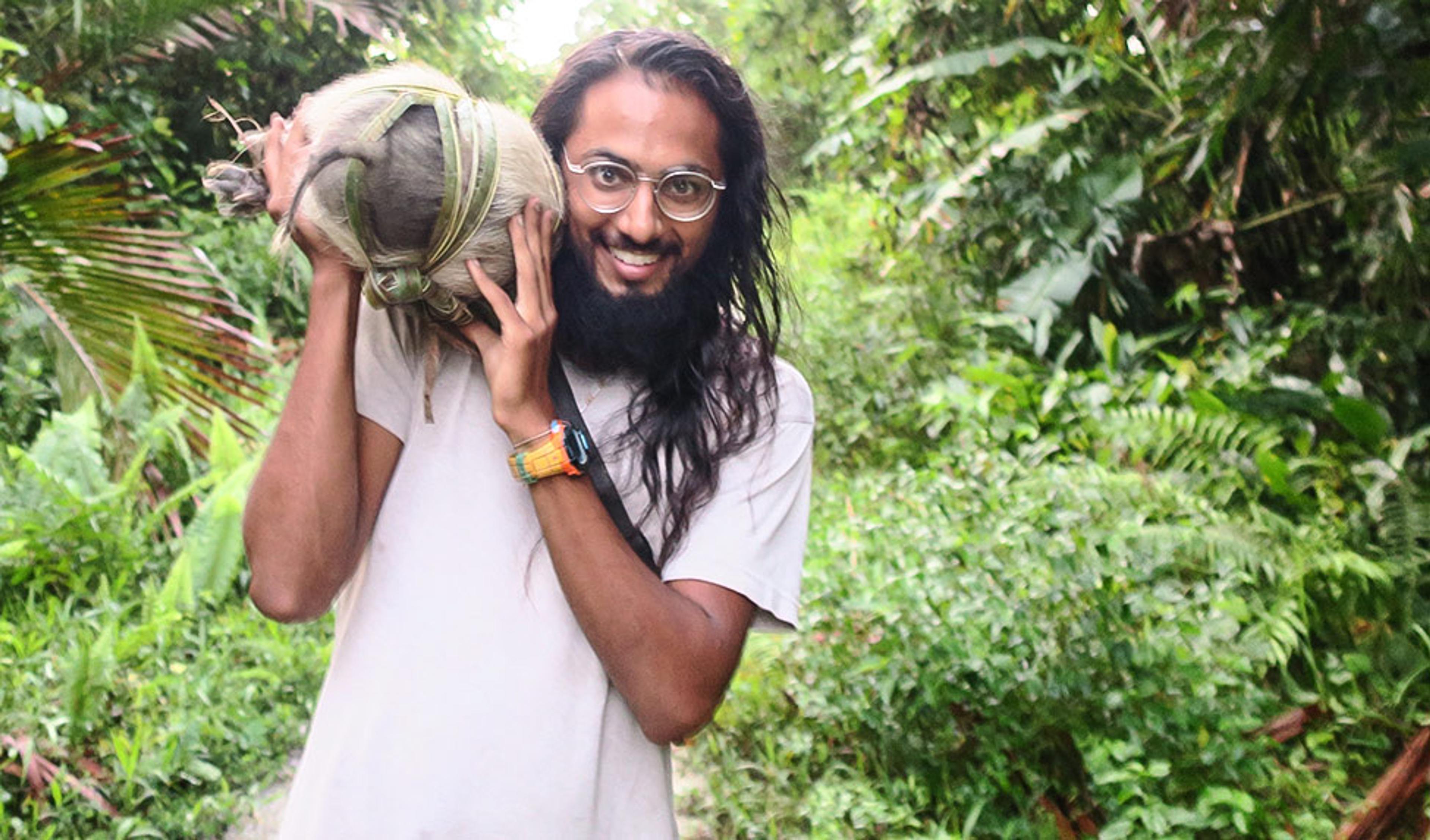 A smiling bearded man with long hair and glasses holding a coconut in a lush, tropical forest setting.