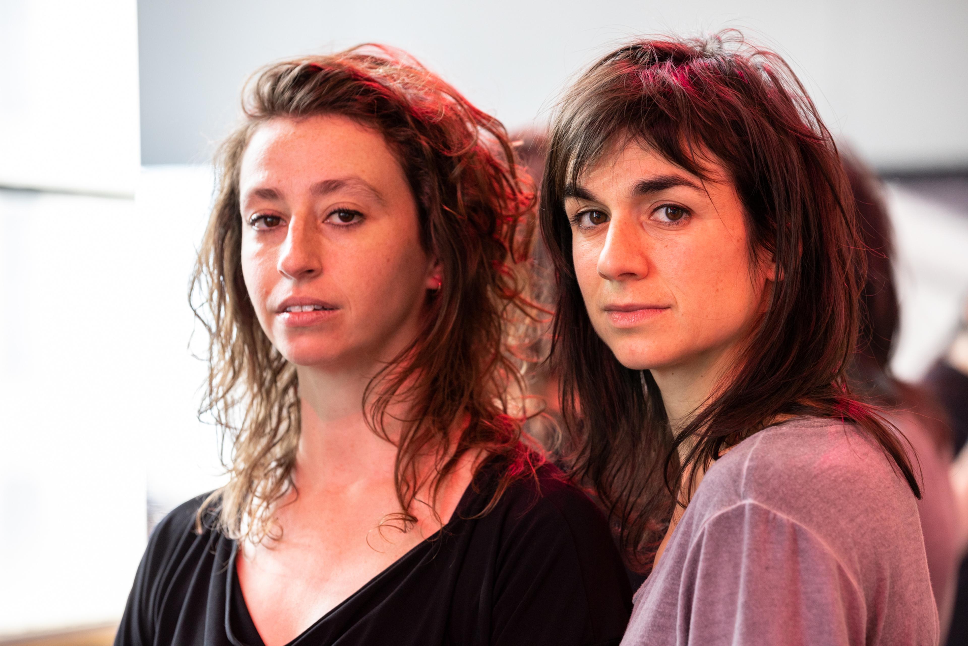 Two women with medium length hair in casual clothing looking at the camera, lit with natural and red light.