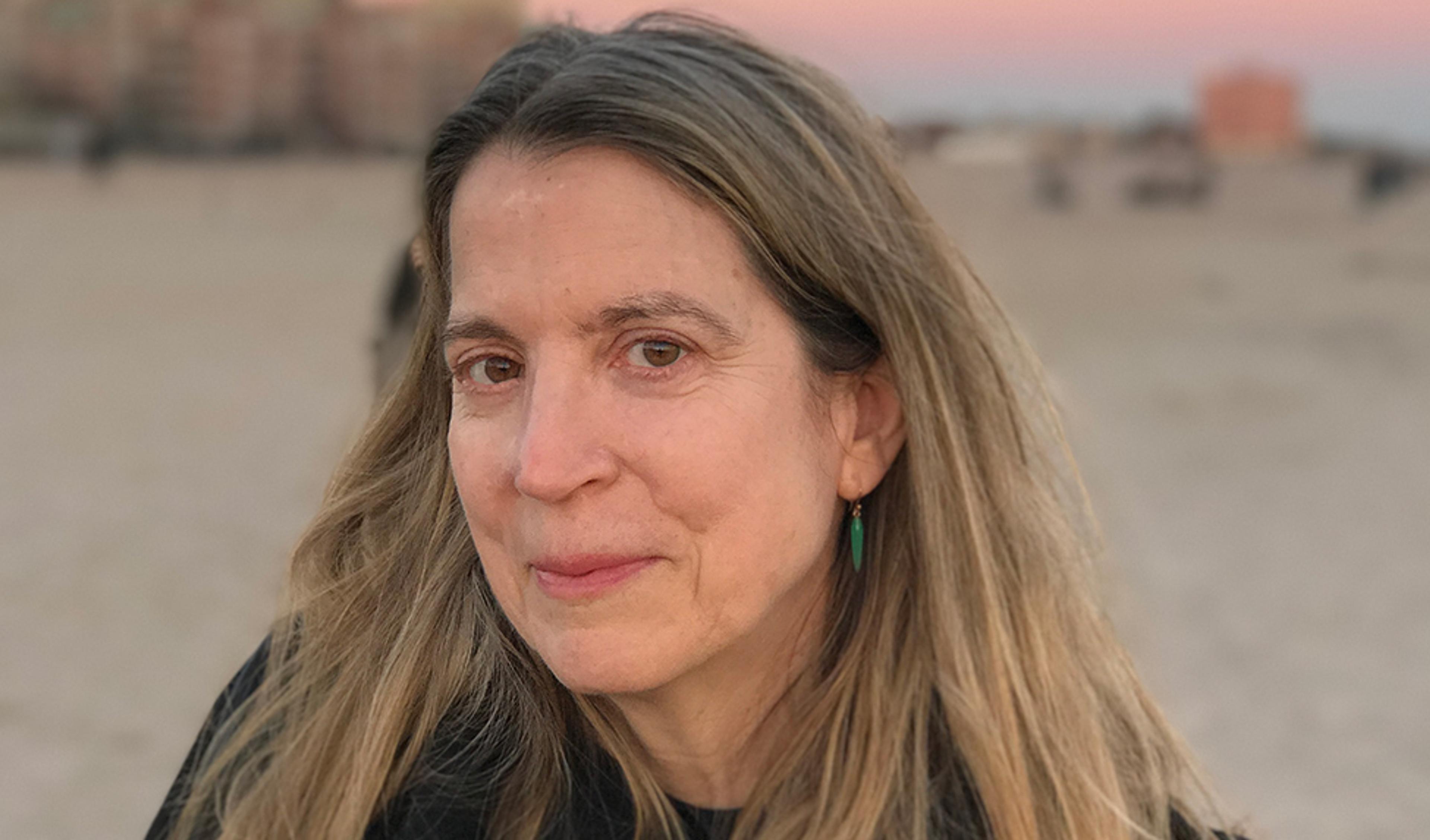 A woman with long hair and green earrings on a beach at sunset, with buildings blurred in the background.