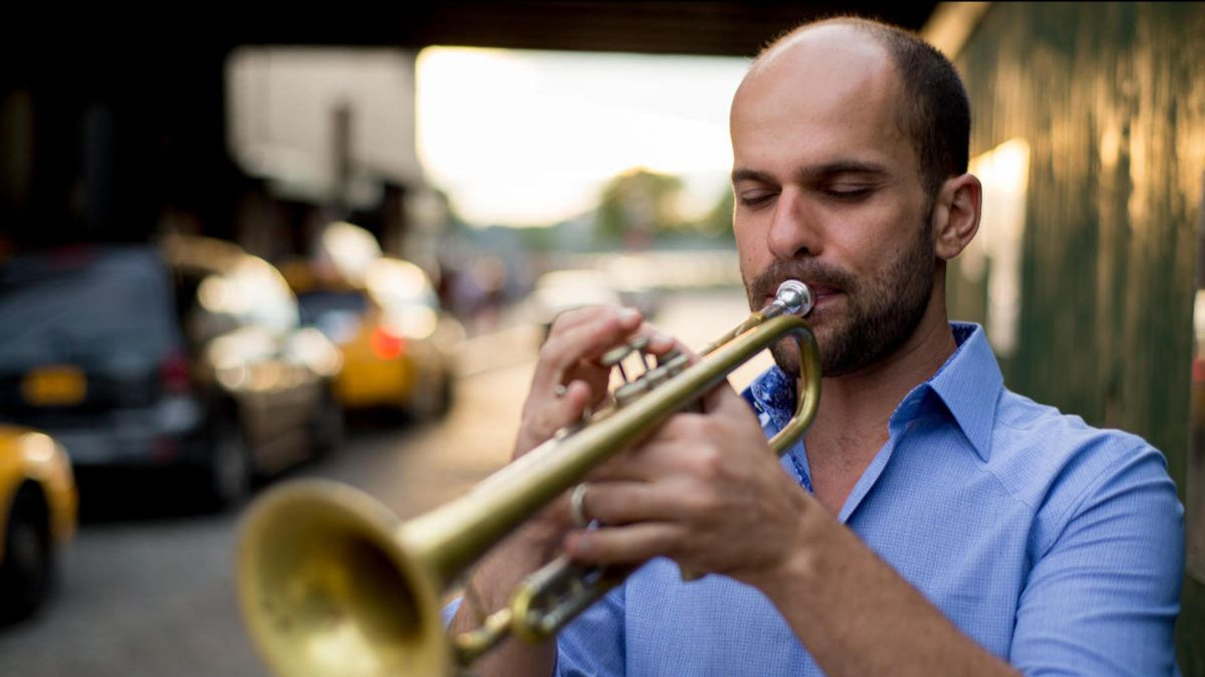 A man in a blue shirt playing a trumpet on a street with blurry cars and buildings in the background.