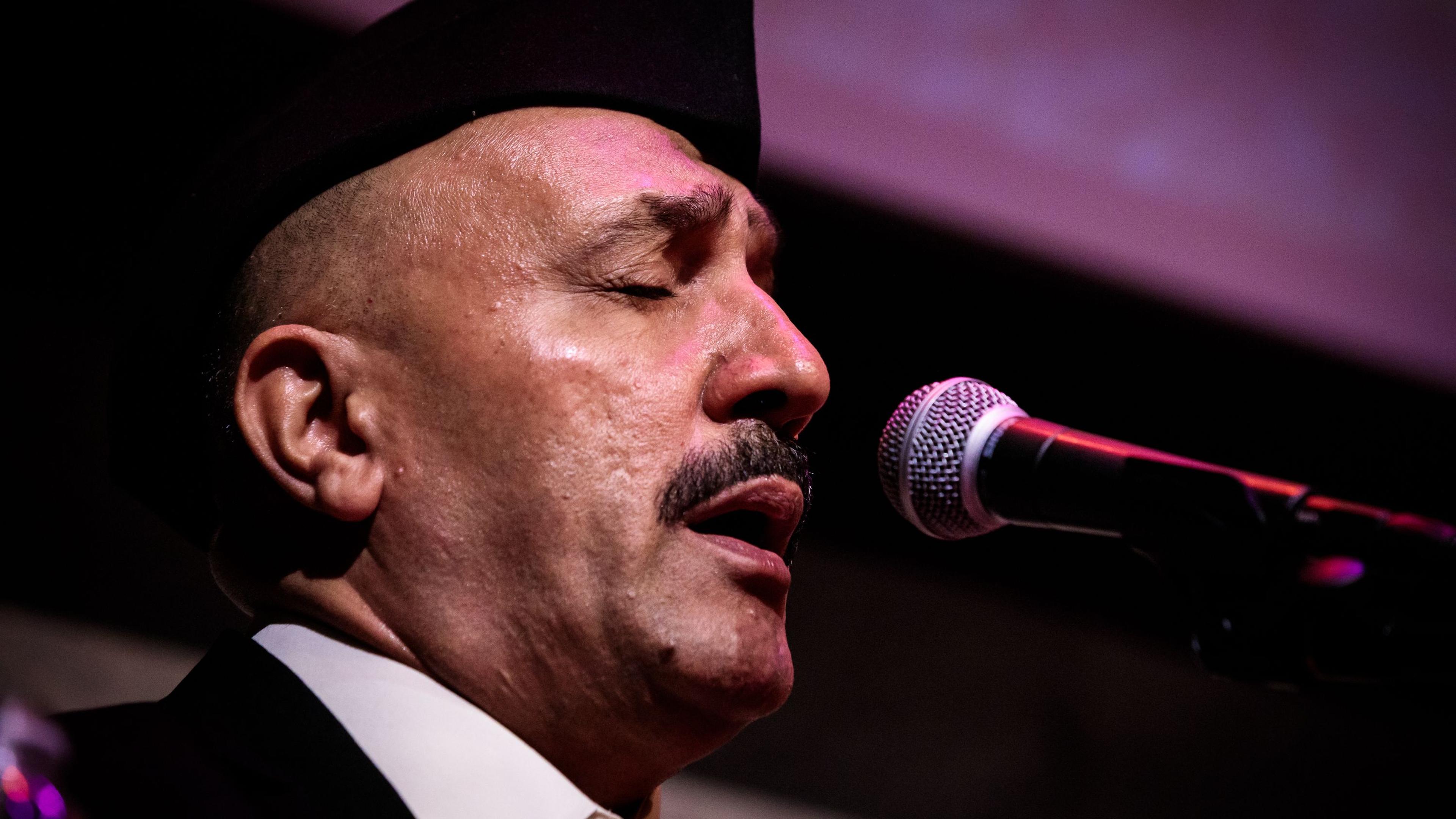 Close-up photo of a man wearing a hat, singing passionately into a microphone with his eyes closed under stage lighting.