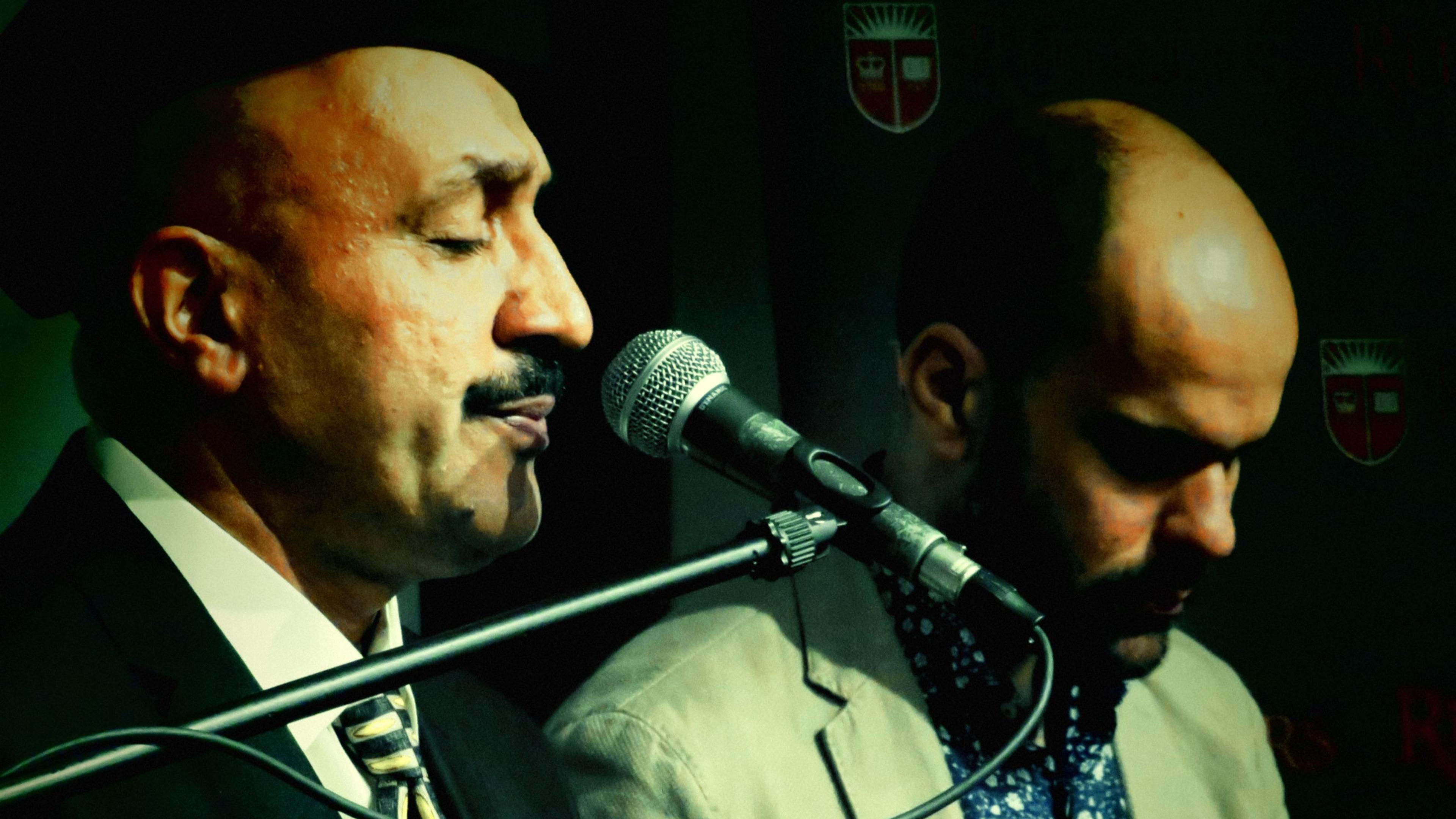 Photo of two men, one in a suit singing into a microphone while the other stands beside him in front of a dark background.