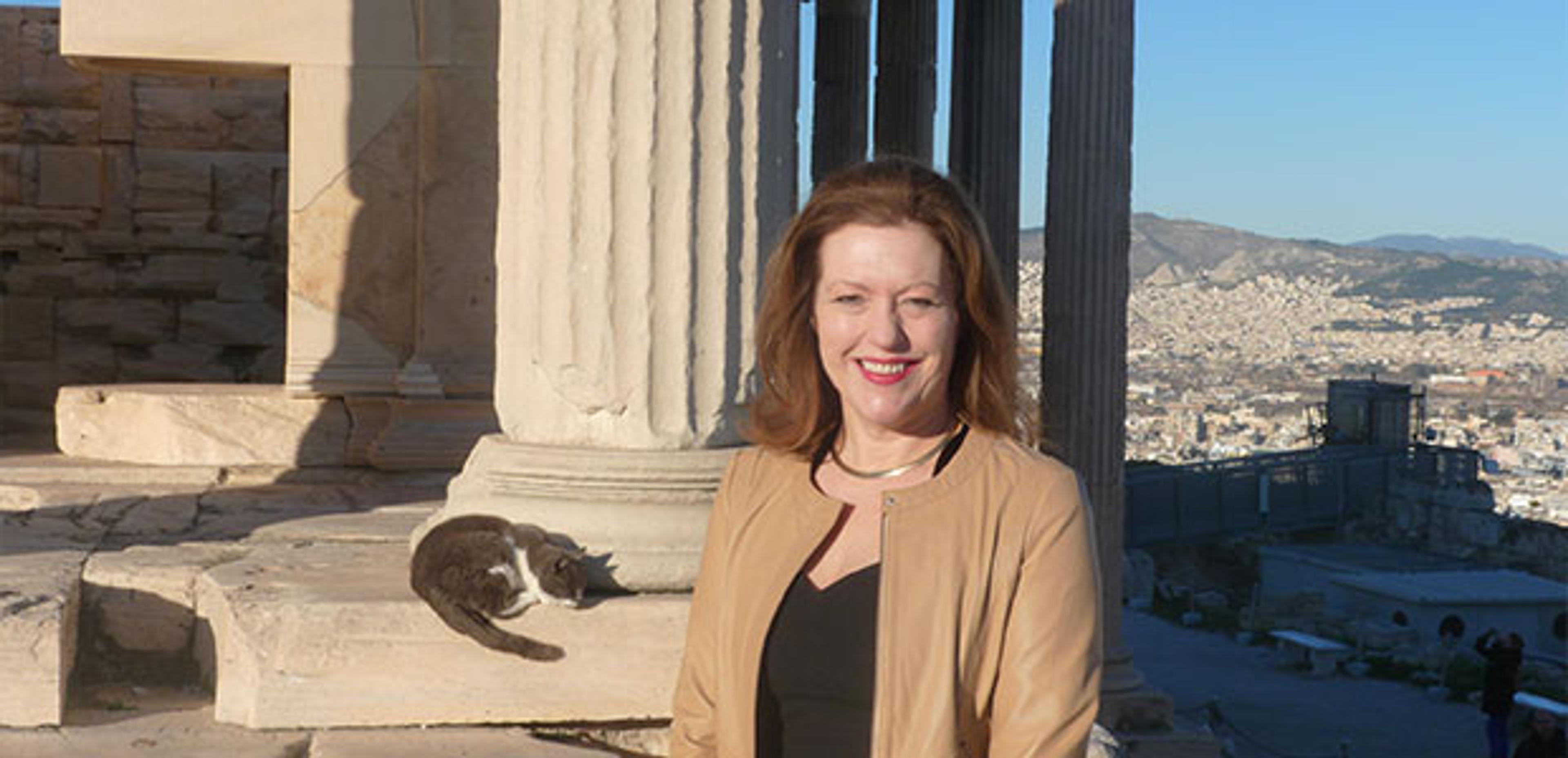 A woman stands in front of ancient ruins with a sleeping cat nearby. Cityscape in the background. Photo taken outdoors in daylight.