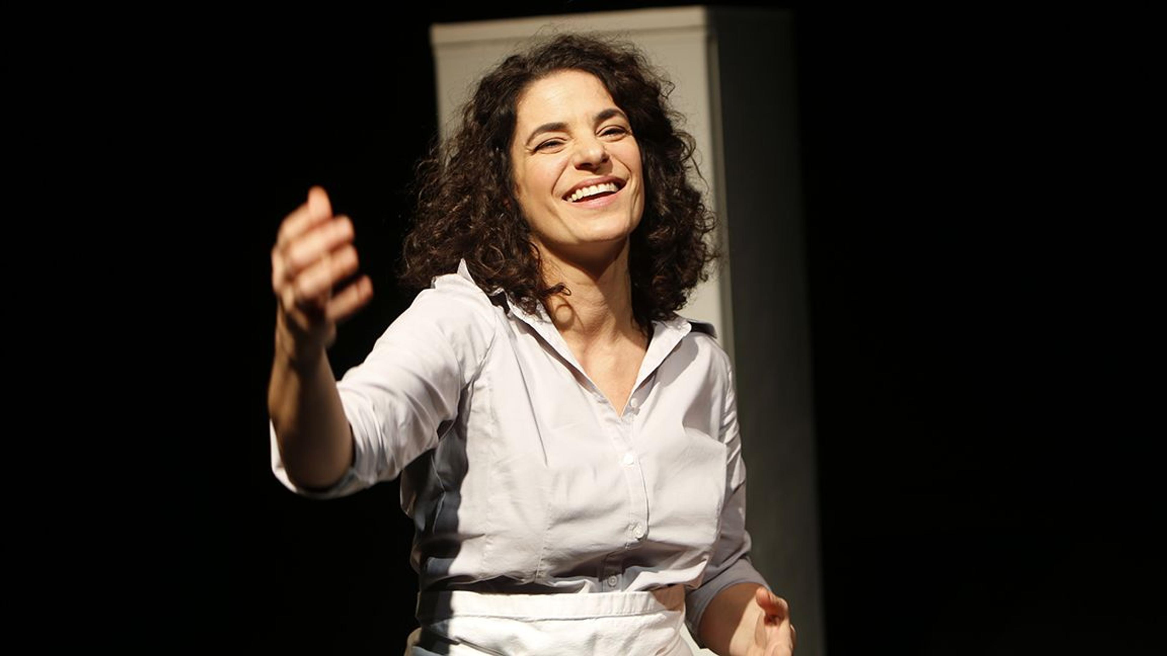 A smiling woman with curly hair wearing a white shirt, reaching out with her right hand, against a dark background.