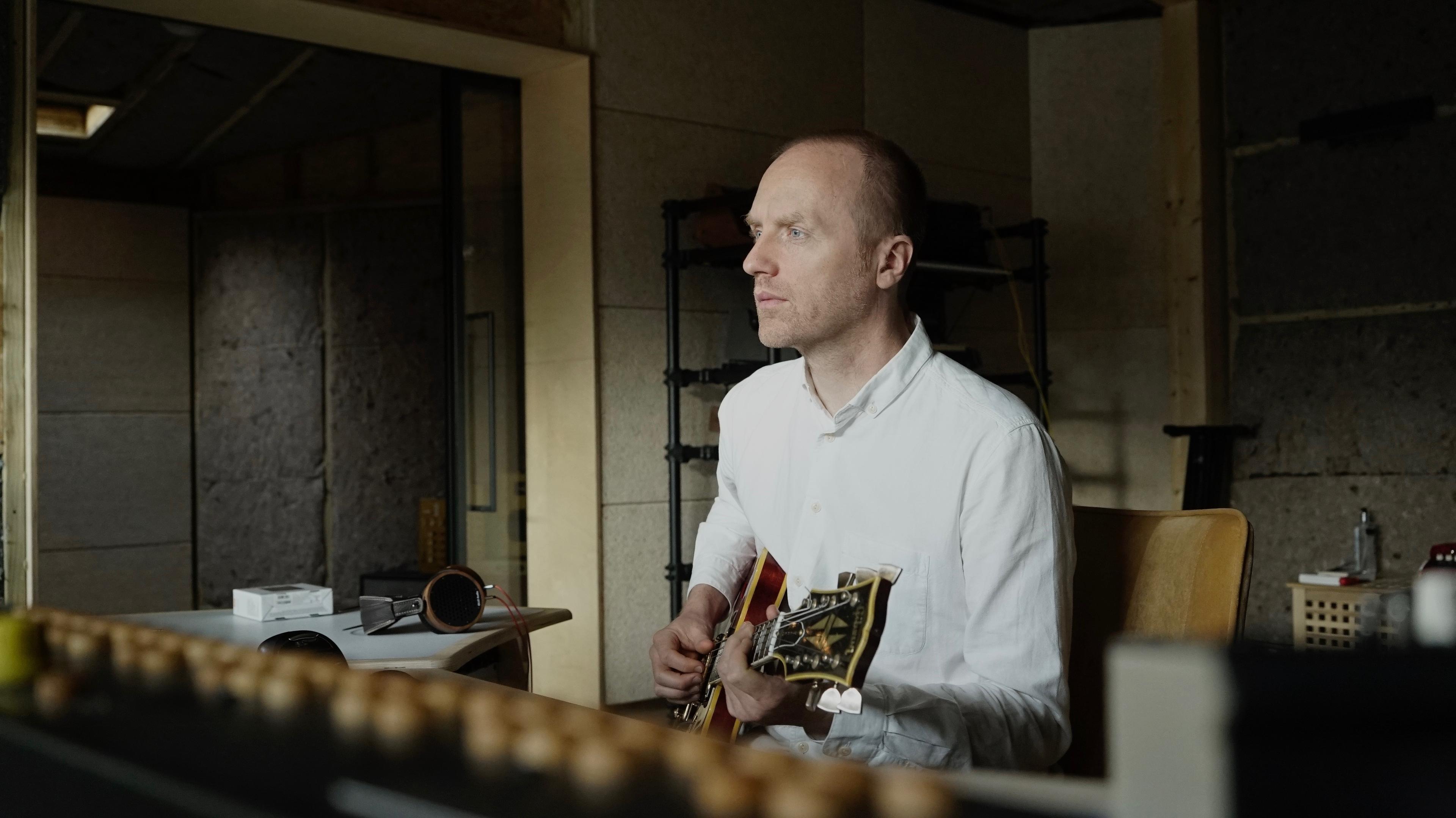 A man in a white shirt playing an electric guitar in a recording studio with a focused expression.