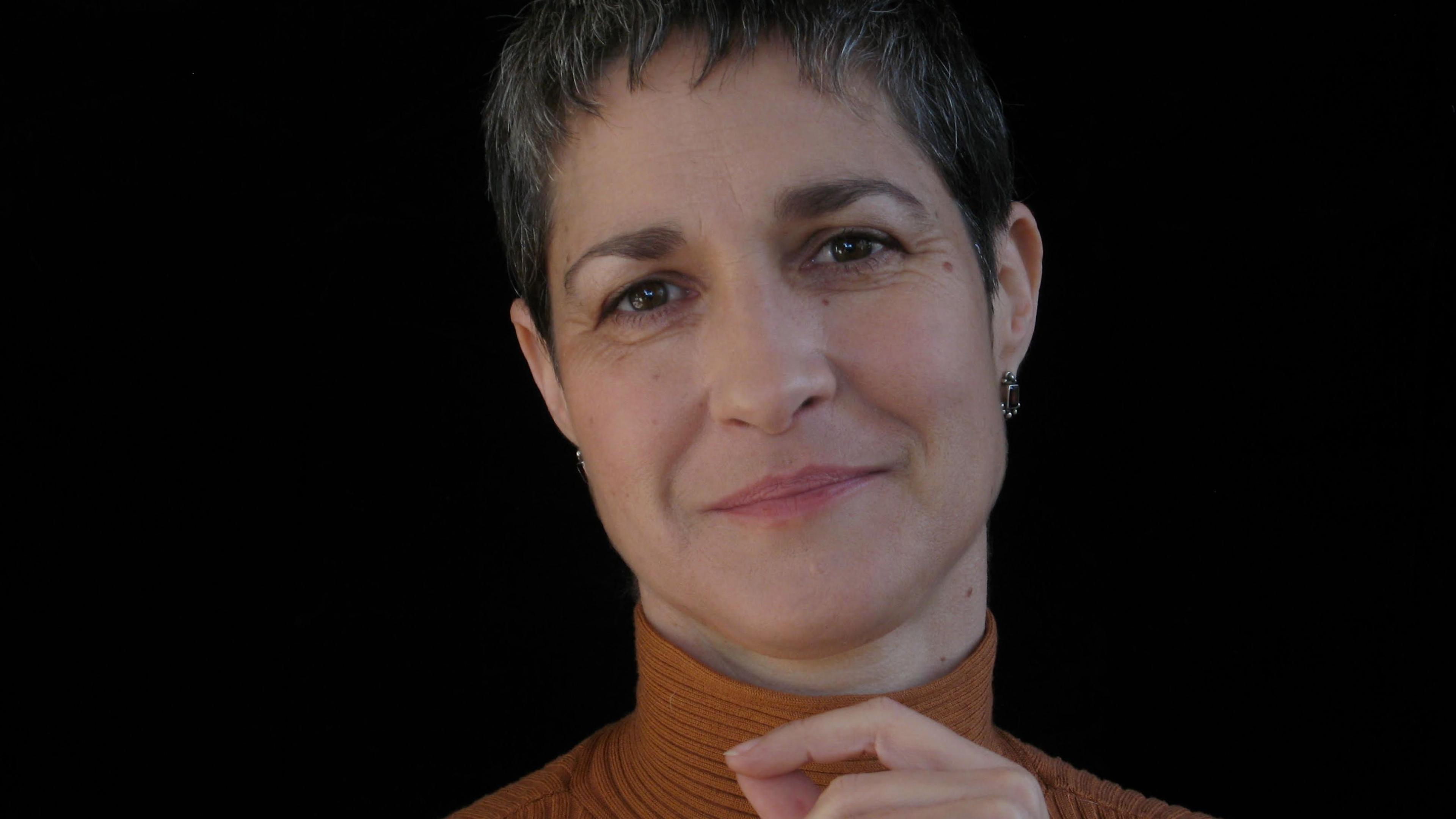 A woman with short grey hair in a brown turtleneck, smiling softly against a black background.