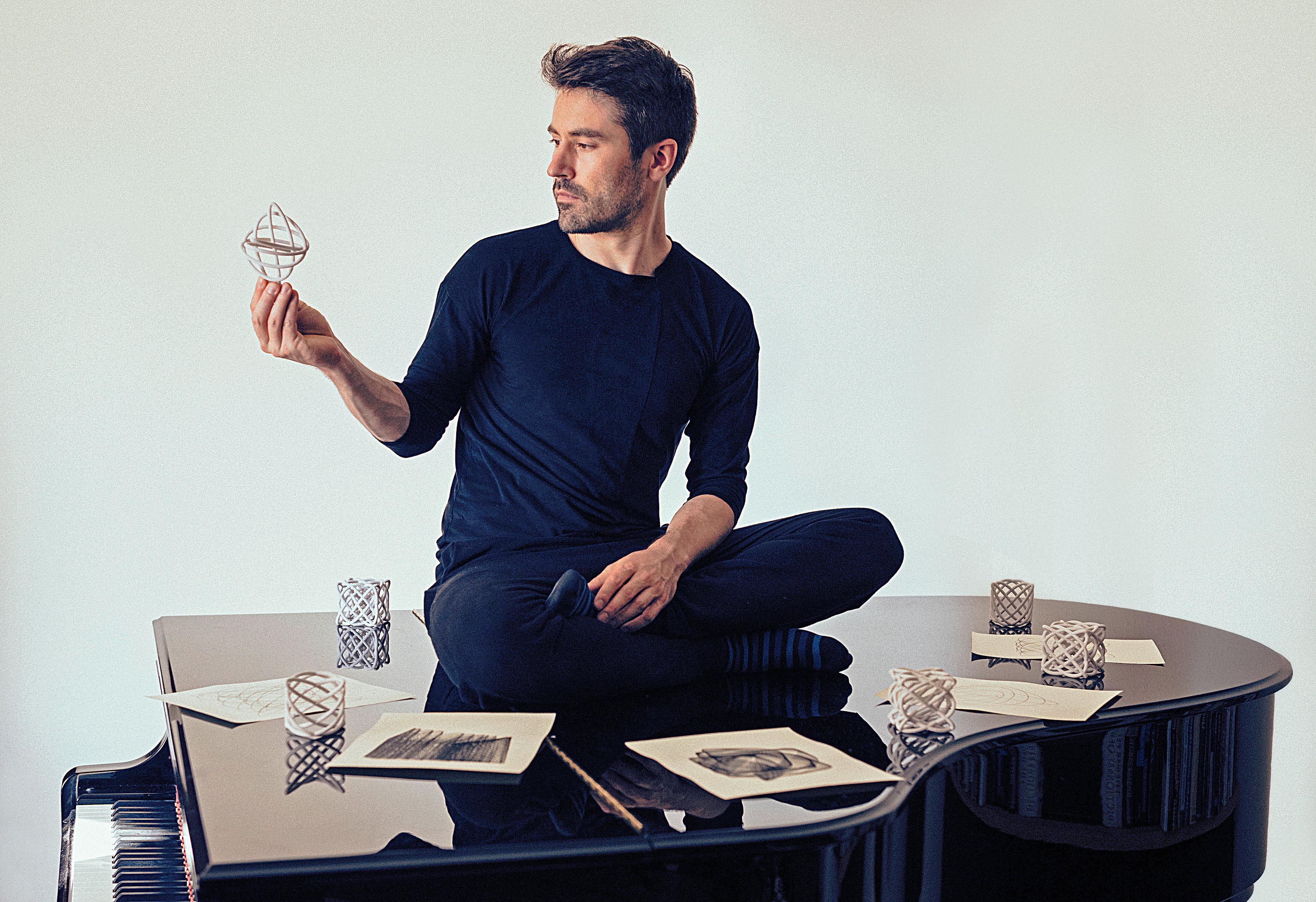 A man in black sitting on a piano, holding a geometric object, surrounded by drawings and similar objects with a blank background.