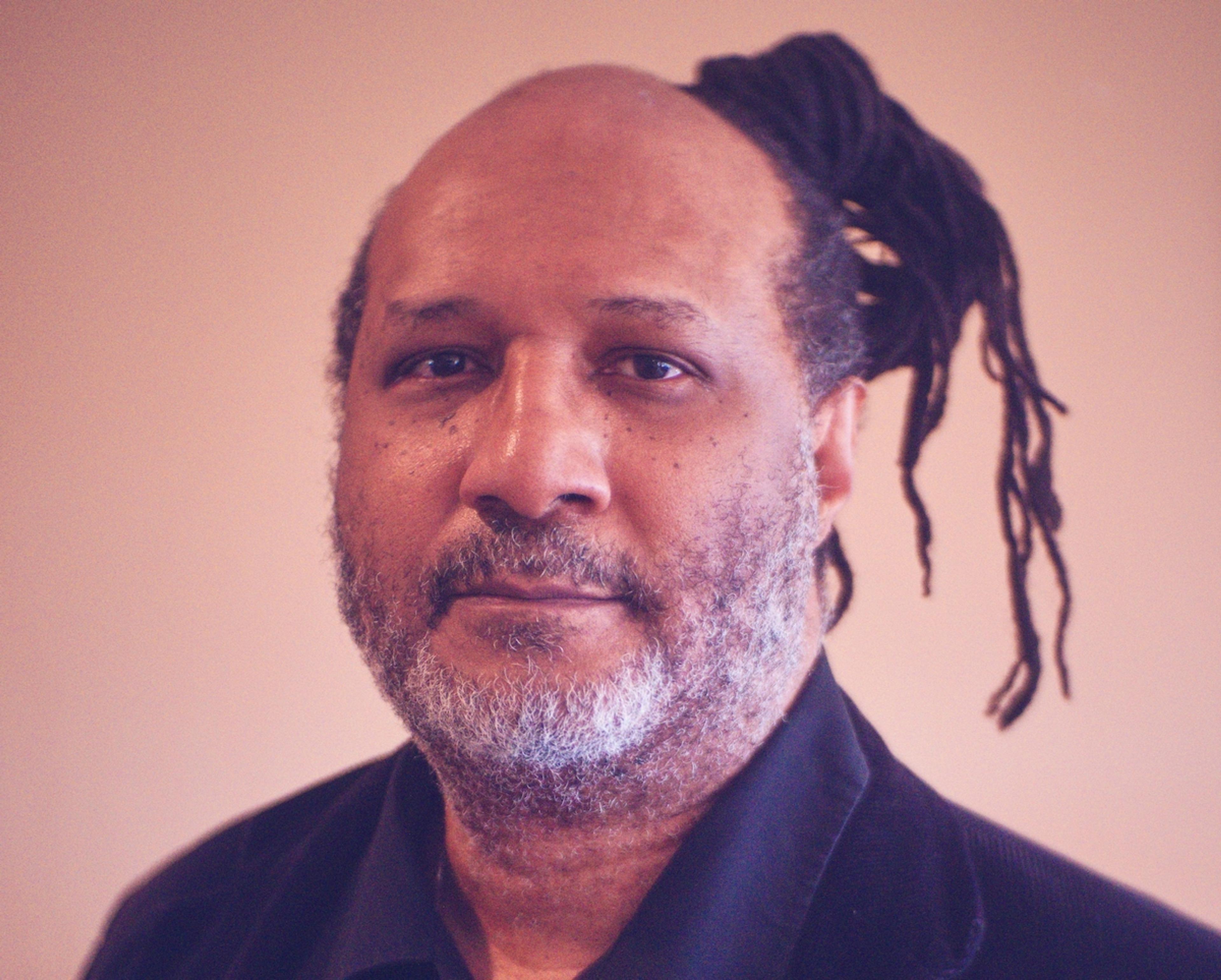 A middle-aged man with a beard and greying hair wearing a black shirt with dreadlocks tied back on a neutral background.