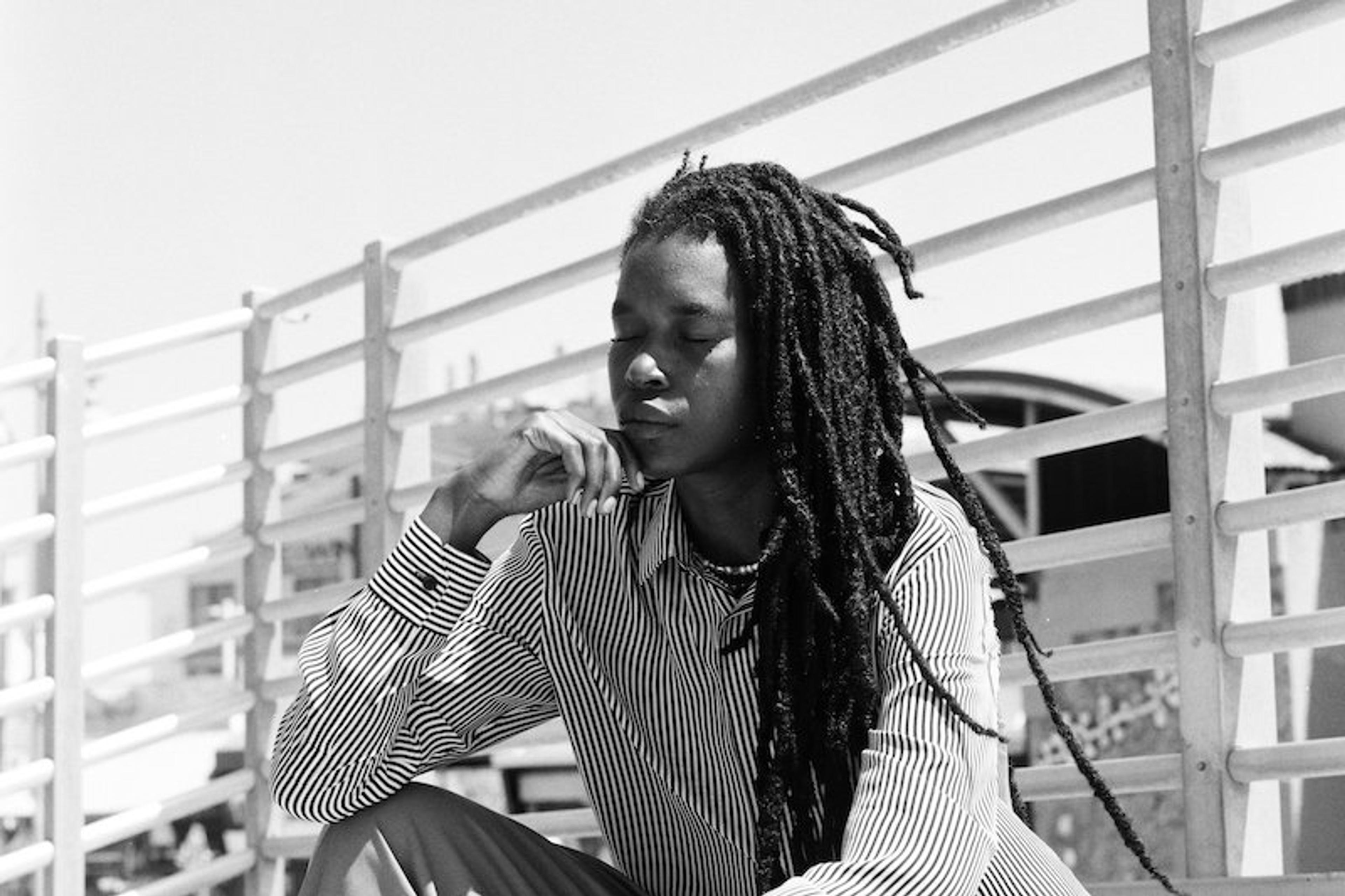 Black person with long hair sitting in front of a railing, wearing a striped shirt. The person appears contemplative.