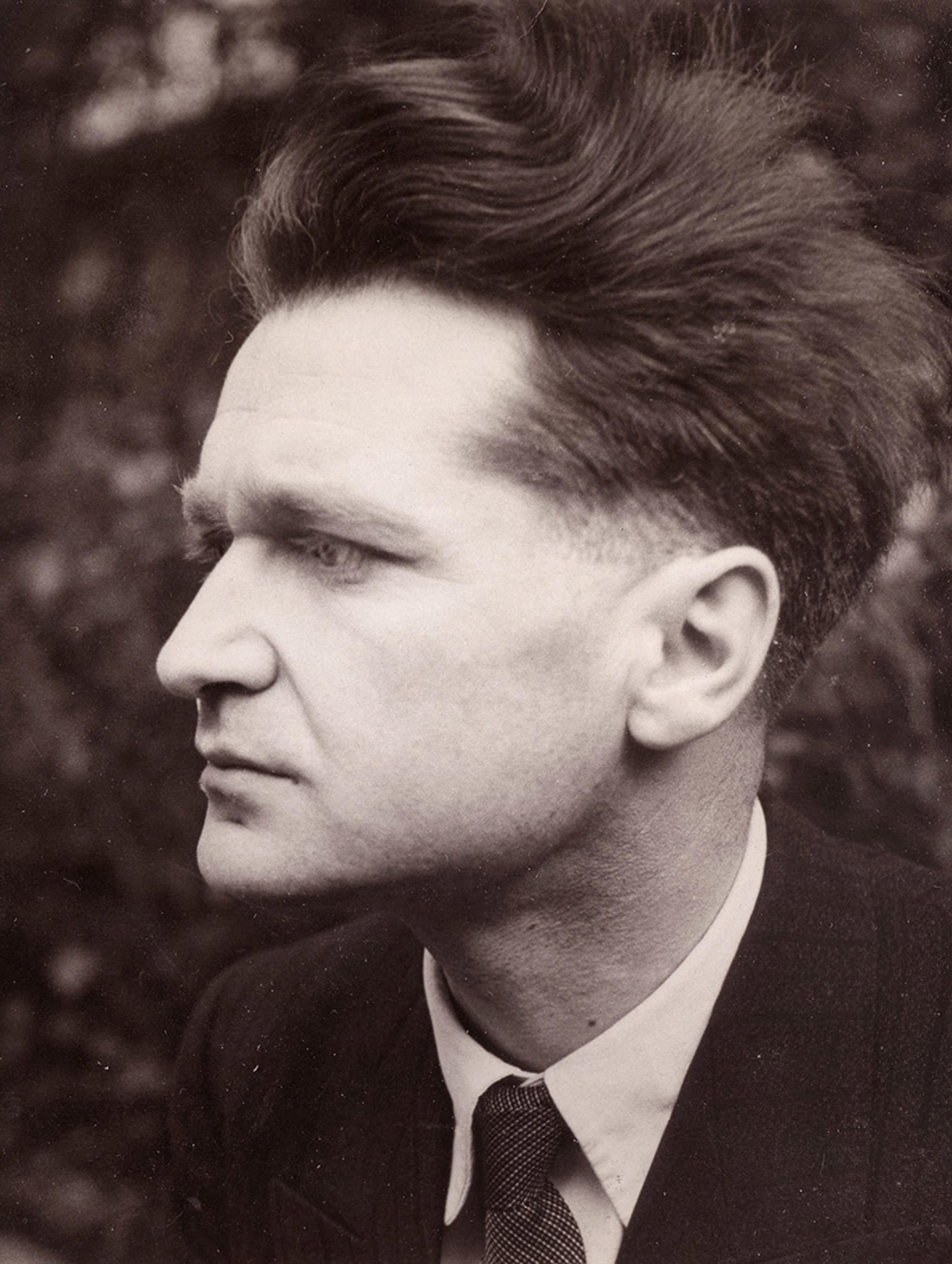 Vintage photo of a man in a suit with a serious expression shot in profile against a blurred outdoor background.