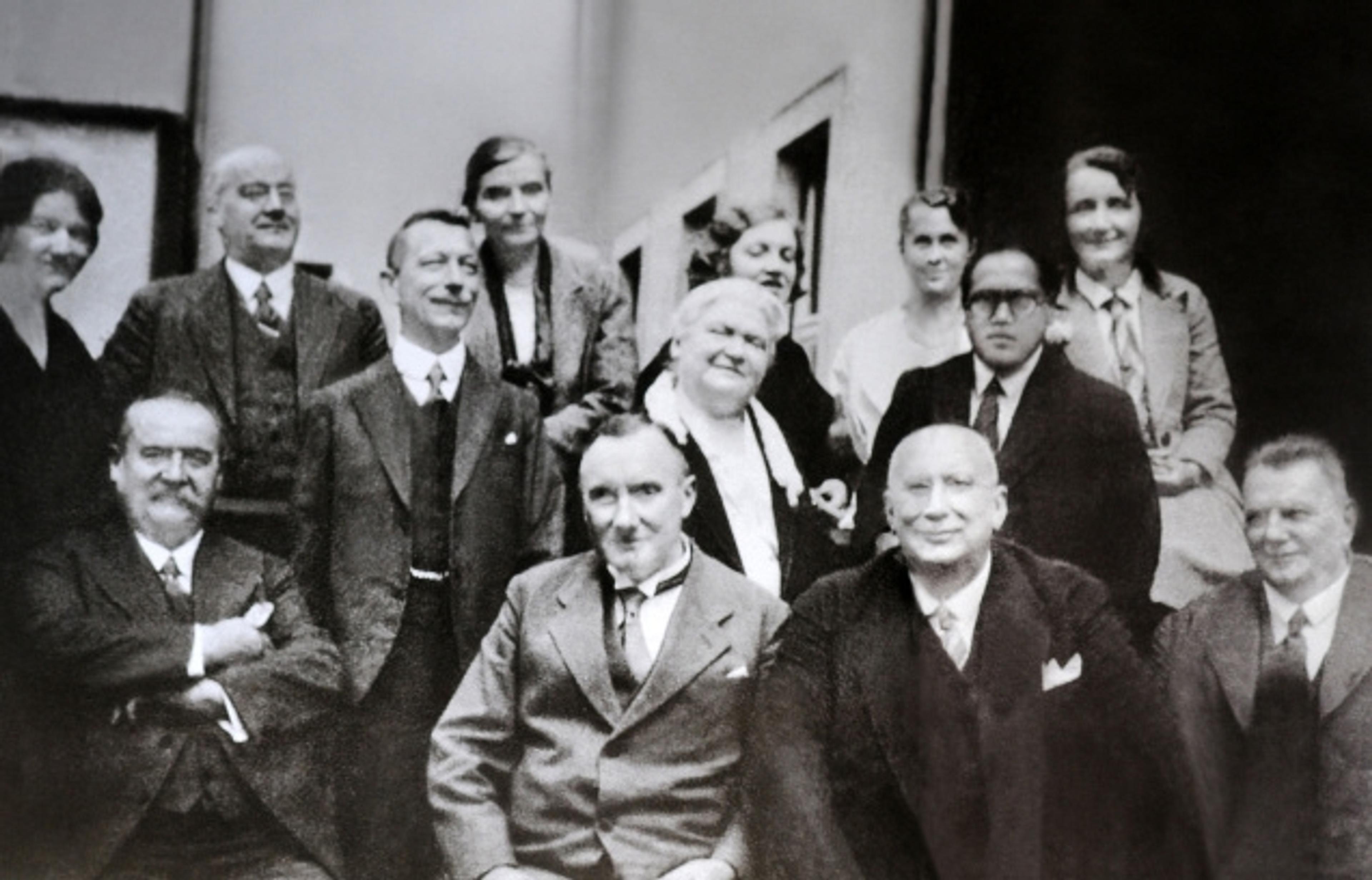 Vintage photo of a group of men and women in formal attire, taken indoors. Some are standing, others seated.