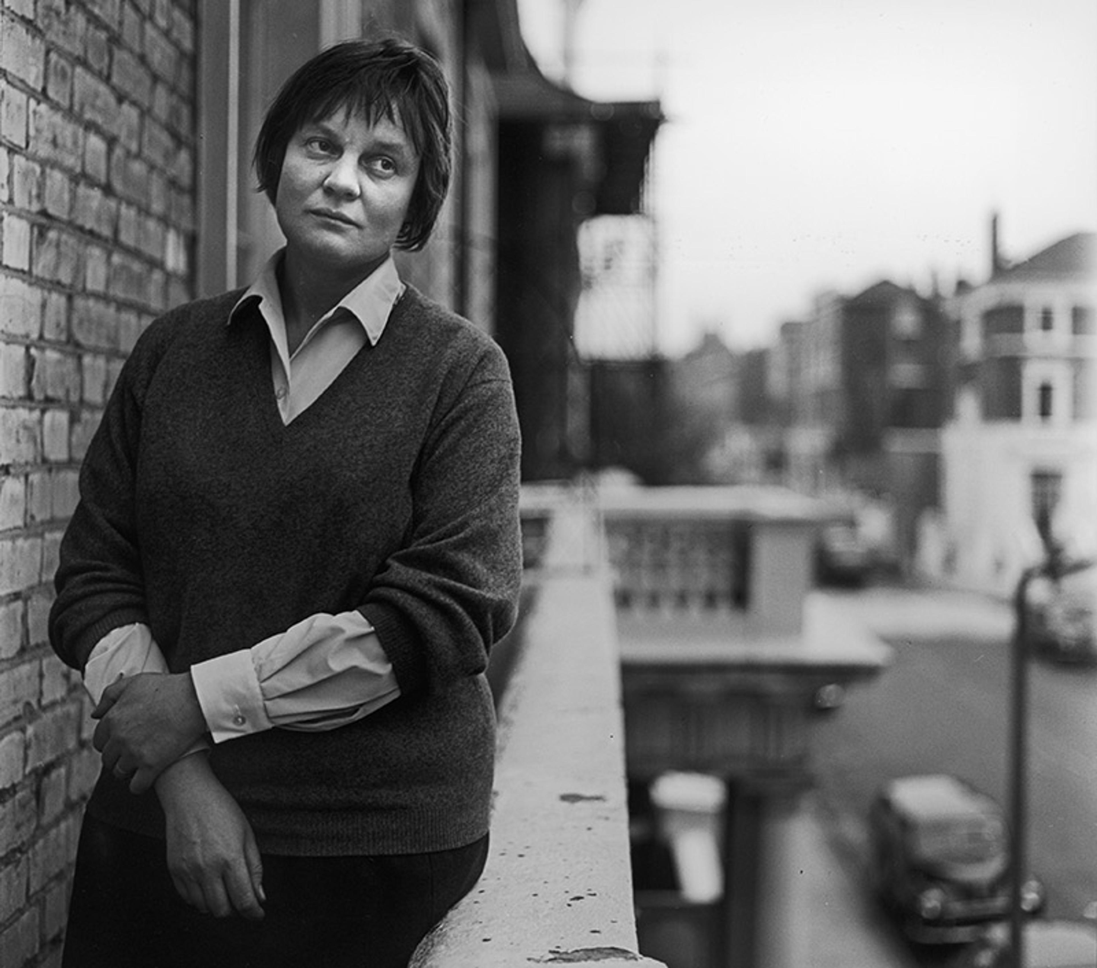 Vintage photo of a woman who is Iris Murdoch leaning on a brick wall on a balcony overlooking a street in an urban area, looking contemplative.