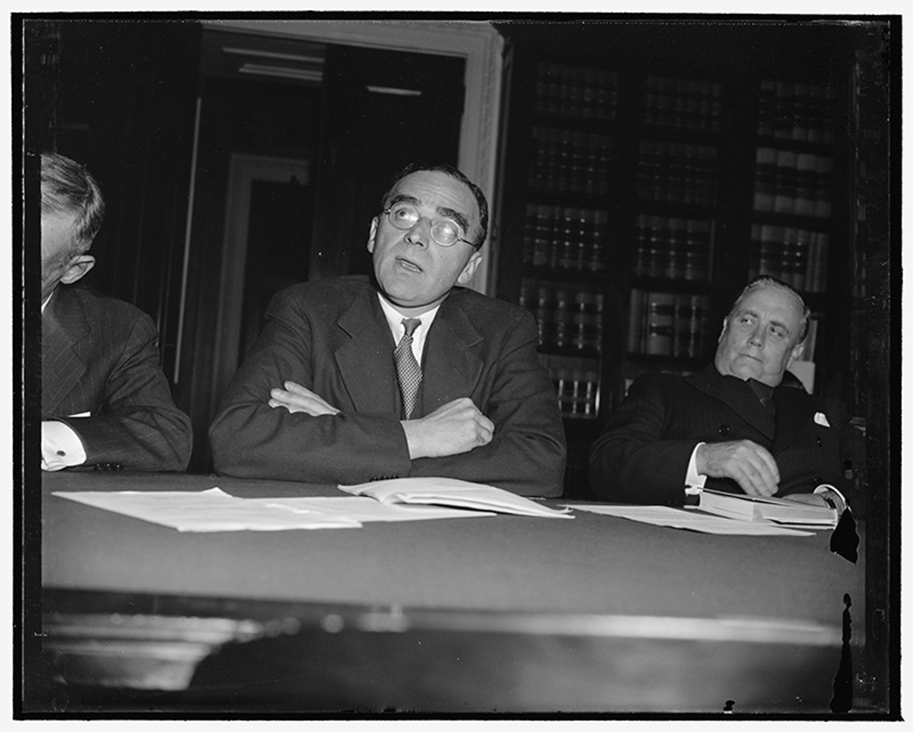 Vintage photo of three men in suits seated at a table with documents in an office. Bookshelves filled with books in the background.