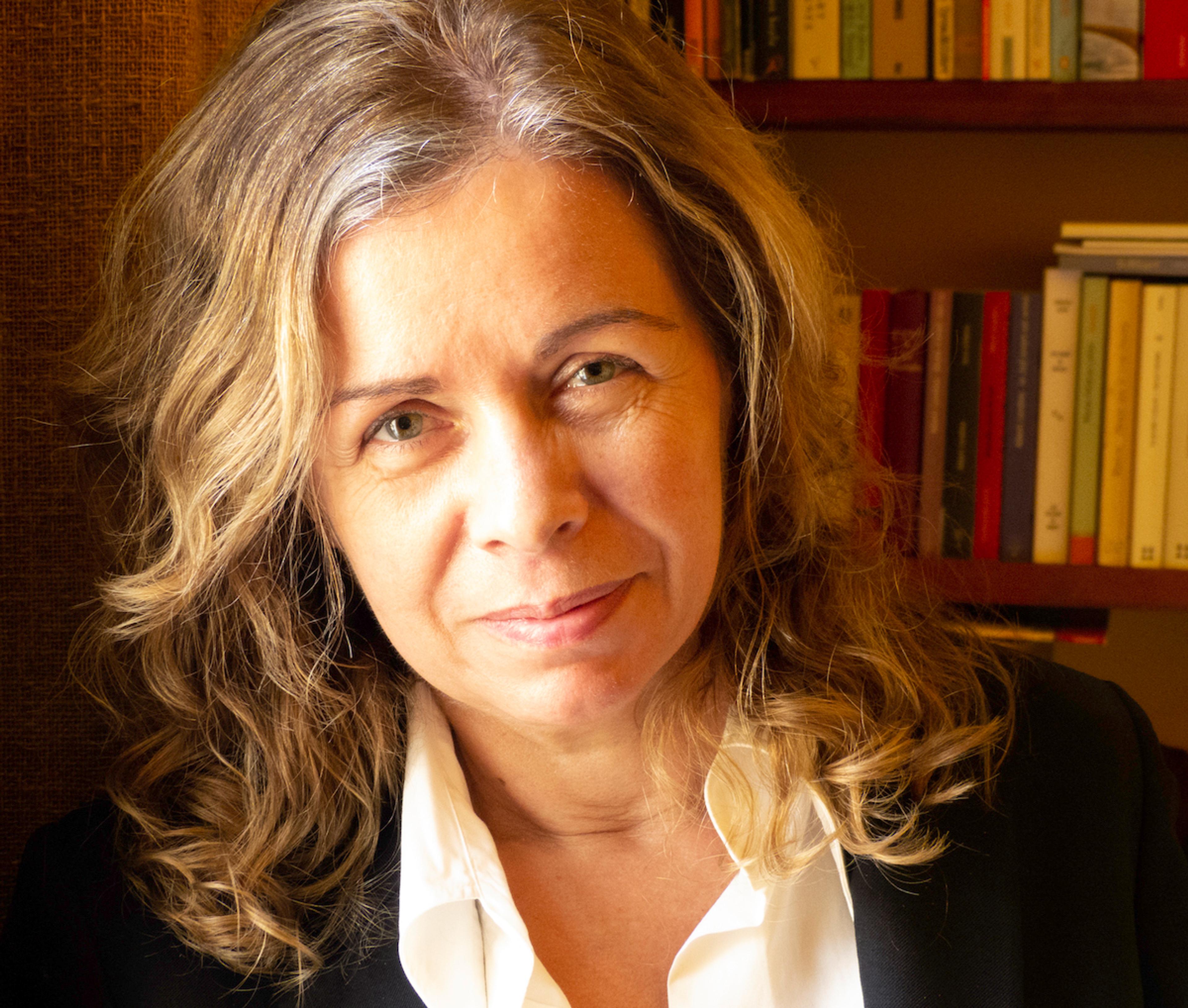 A woman with wavy, shoulder-length hair, wearing a white shirt and black jacket, smiling slightly with bookshelves in the background.