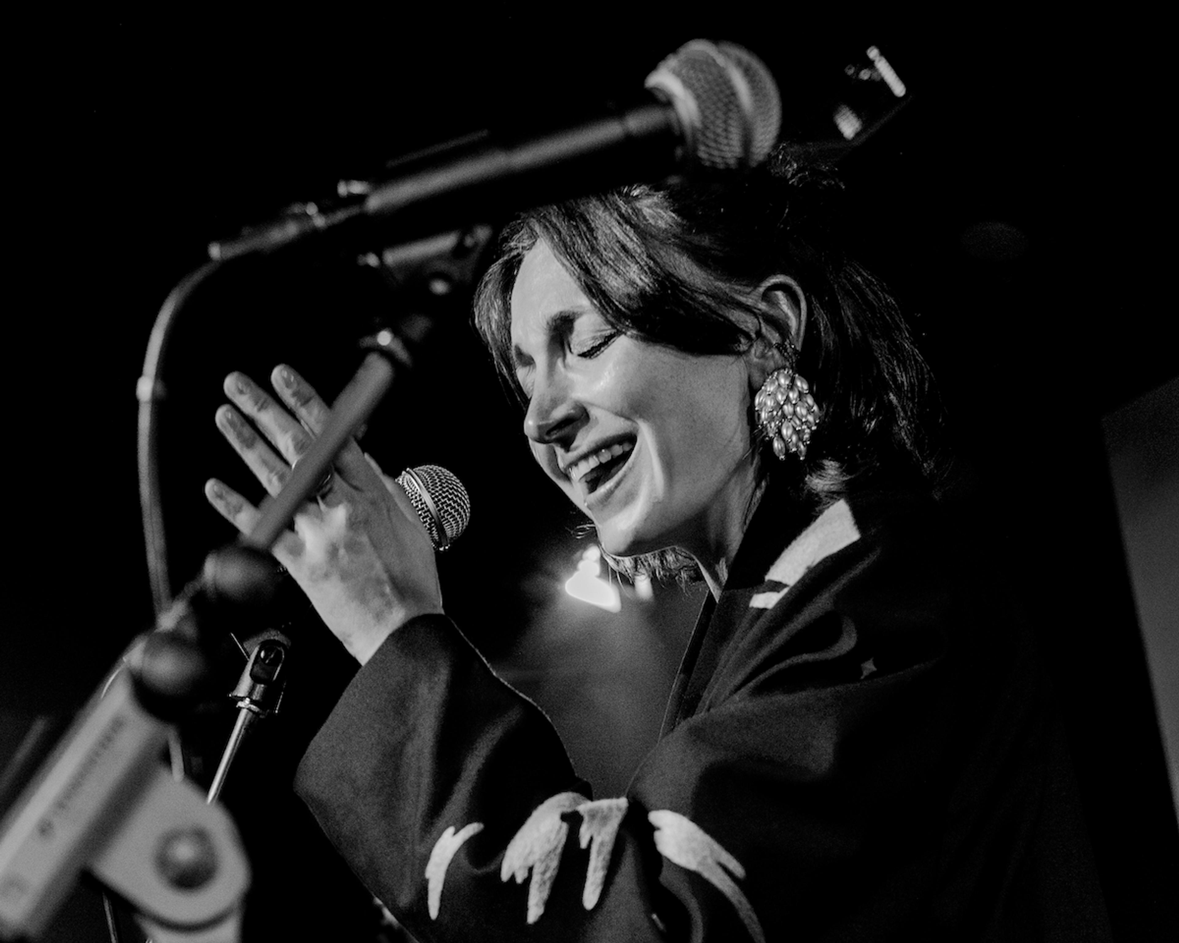 Black-and-white photo of a woman singing passionately into a microphone on stage, with her hands raised and eyes closed.