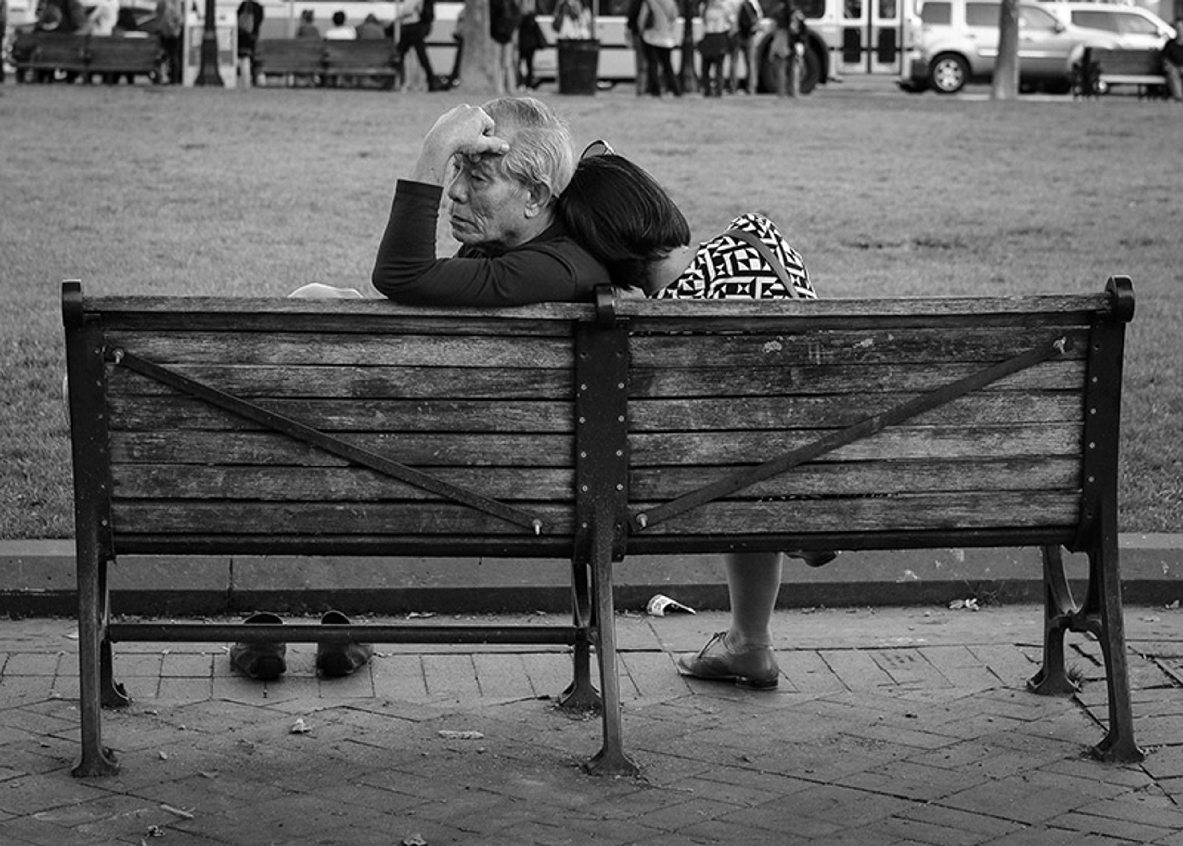 Black-and-white photo of an elderly couple sitting on a wooden bench in a park. The woman leans on the man, who holds his head.