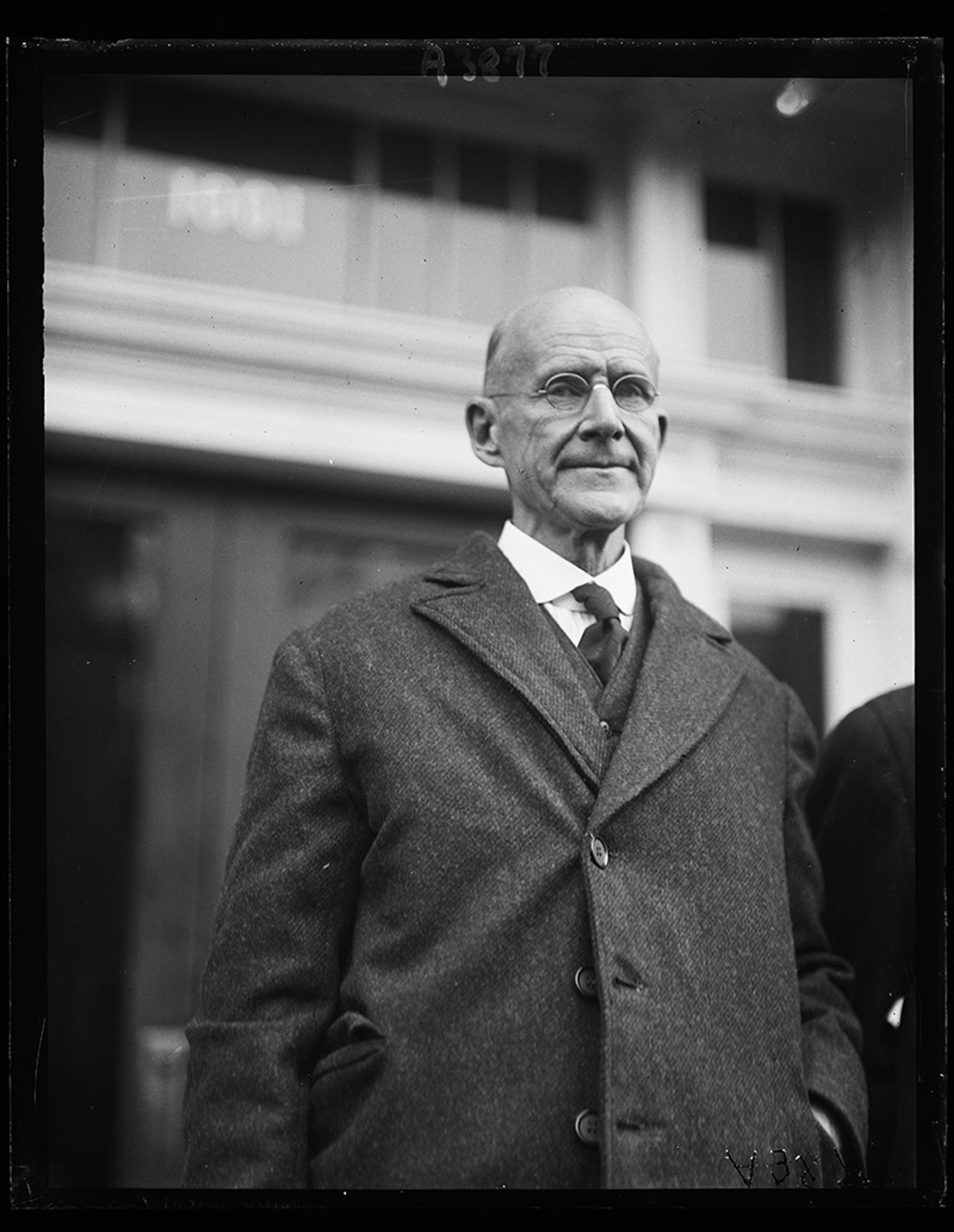 Vintage photo of a bald man wearing a coat, tie and glasses, standing in front of a building with large windows.