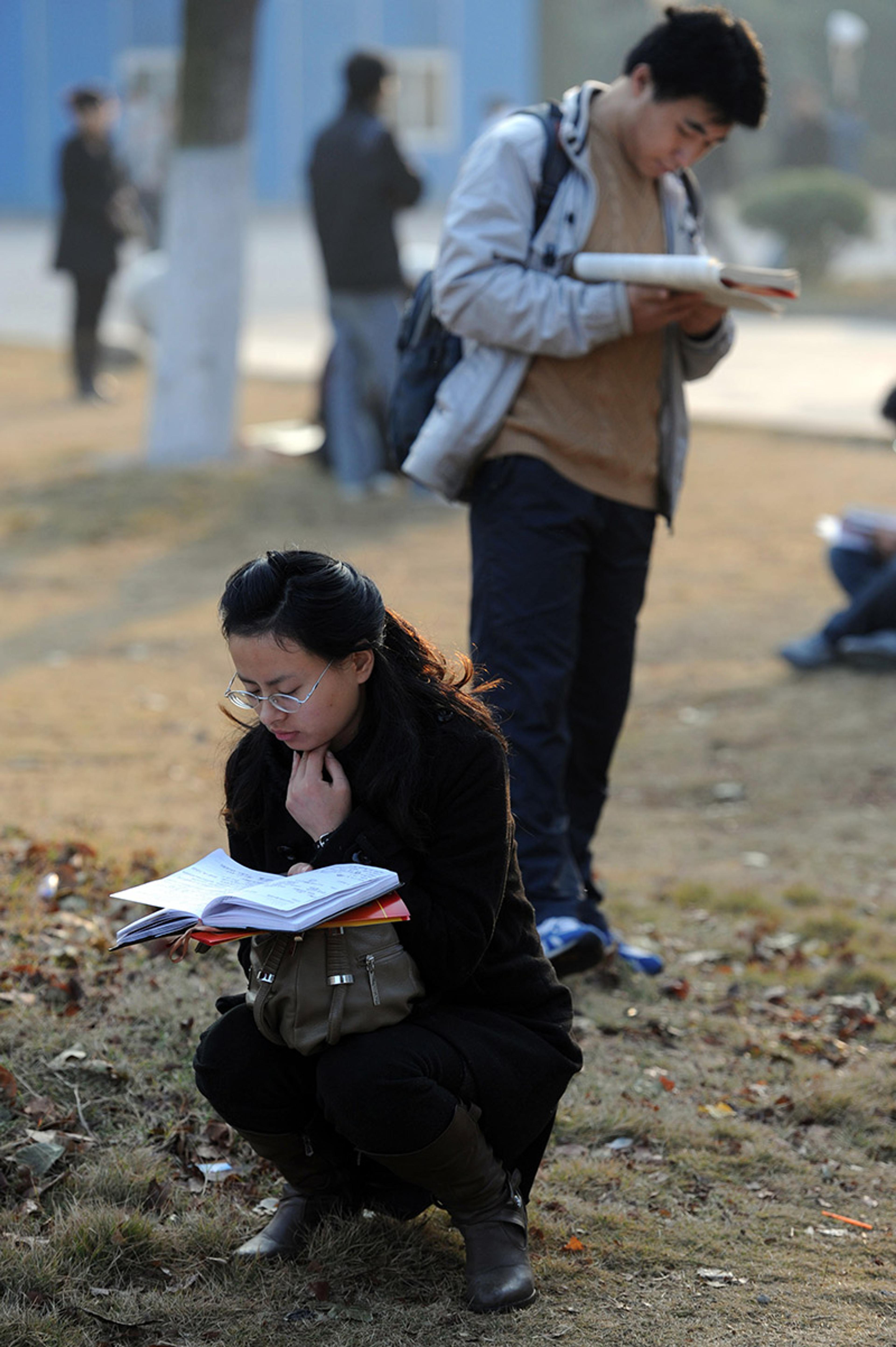 An anxious looking young woman and a young man are reading notebooks or textbooks outside an examination hall