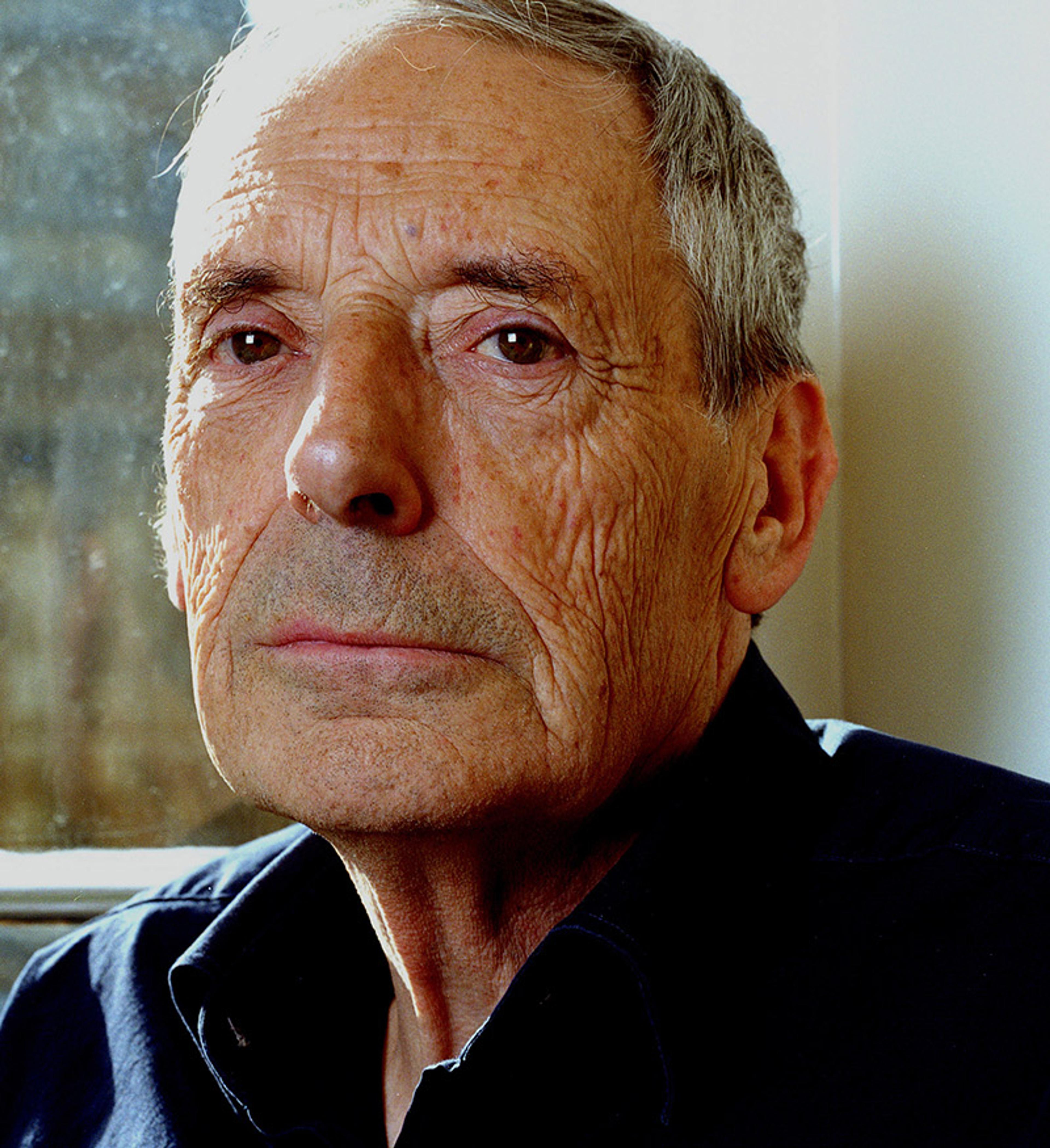 An elderly man with a serious expression, short grey hair and a black shirt, sitting indoors near a window with soft light.