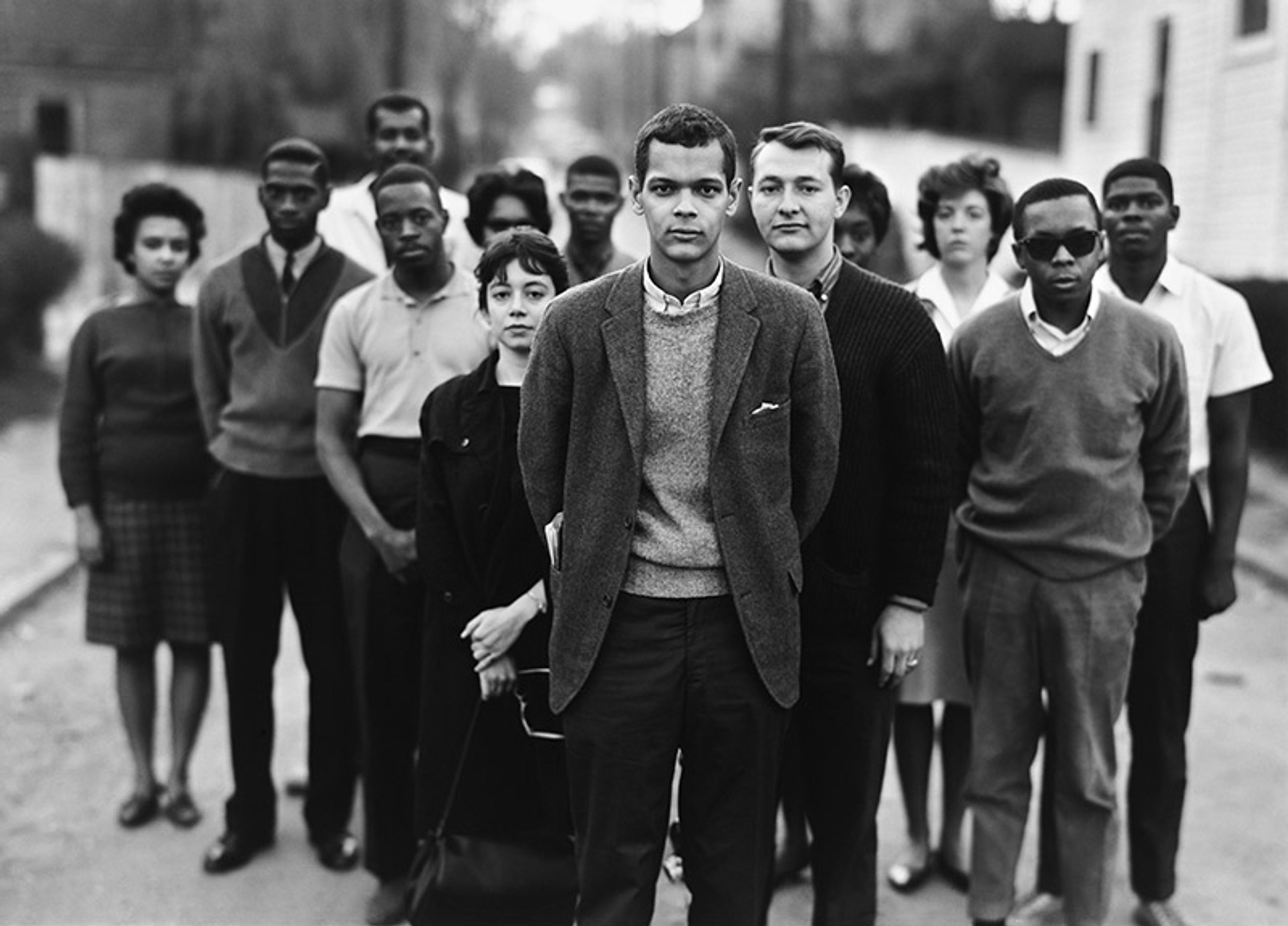 Black-and-white photo of a diverse group of young people standing in a line on an urban street, with one person in the foreground.