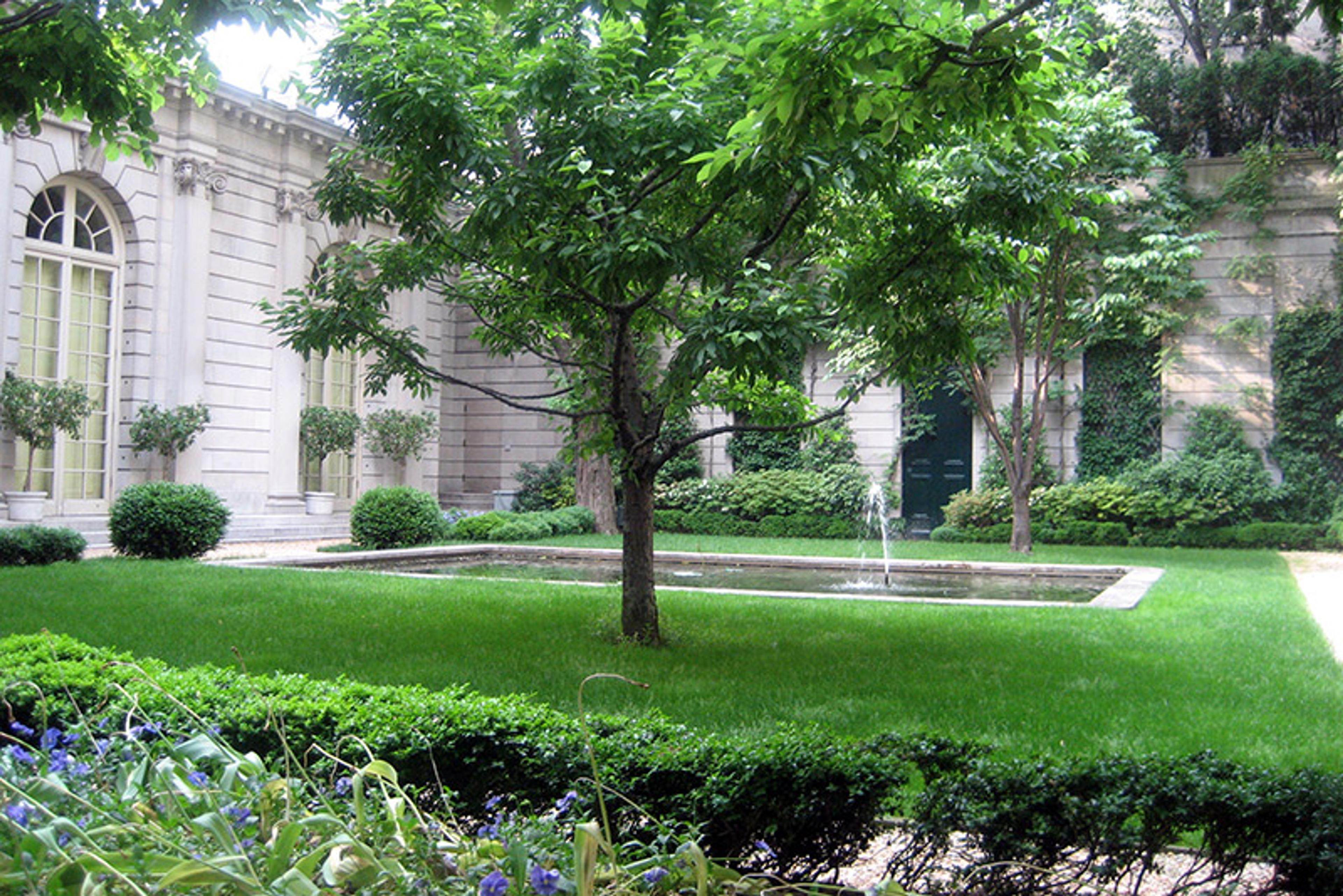 A lush garden with trees, neatly trimmed bushes and a fountain, surrounded by a classical building with arched windows.