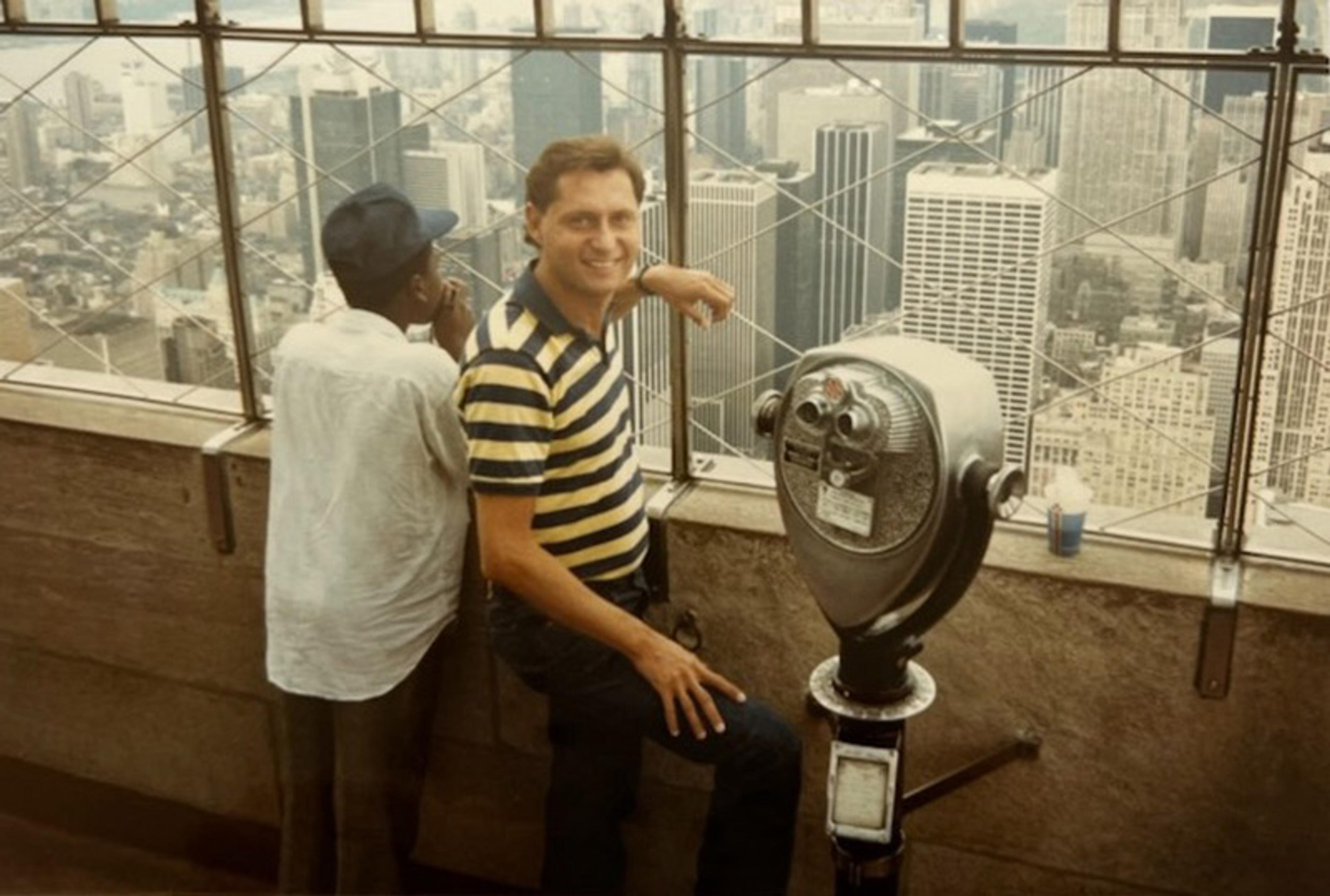 A friendly looking man looks at the camera from a high vantage point above a view of downtown New York City