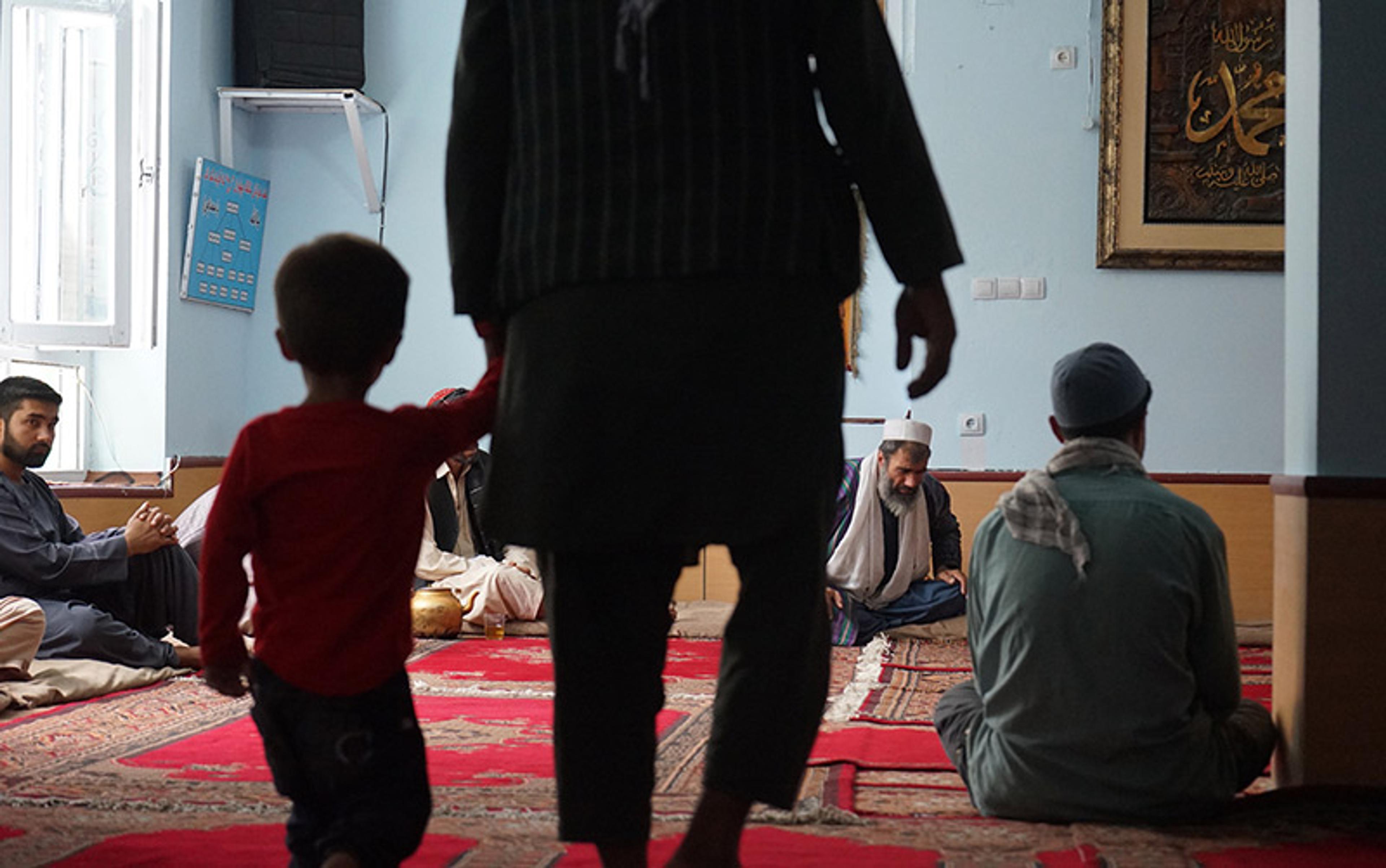 Men and children are seated on the floor of a simple room covered with traditional Afghan carpets