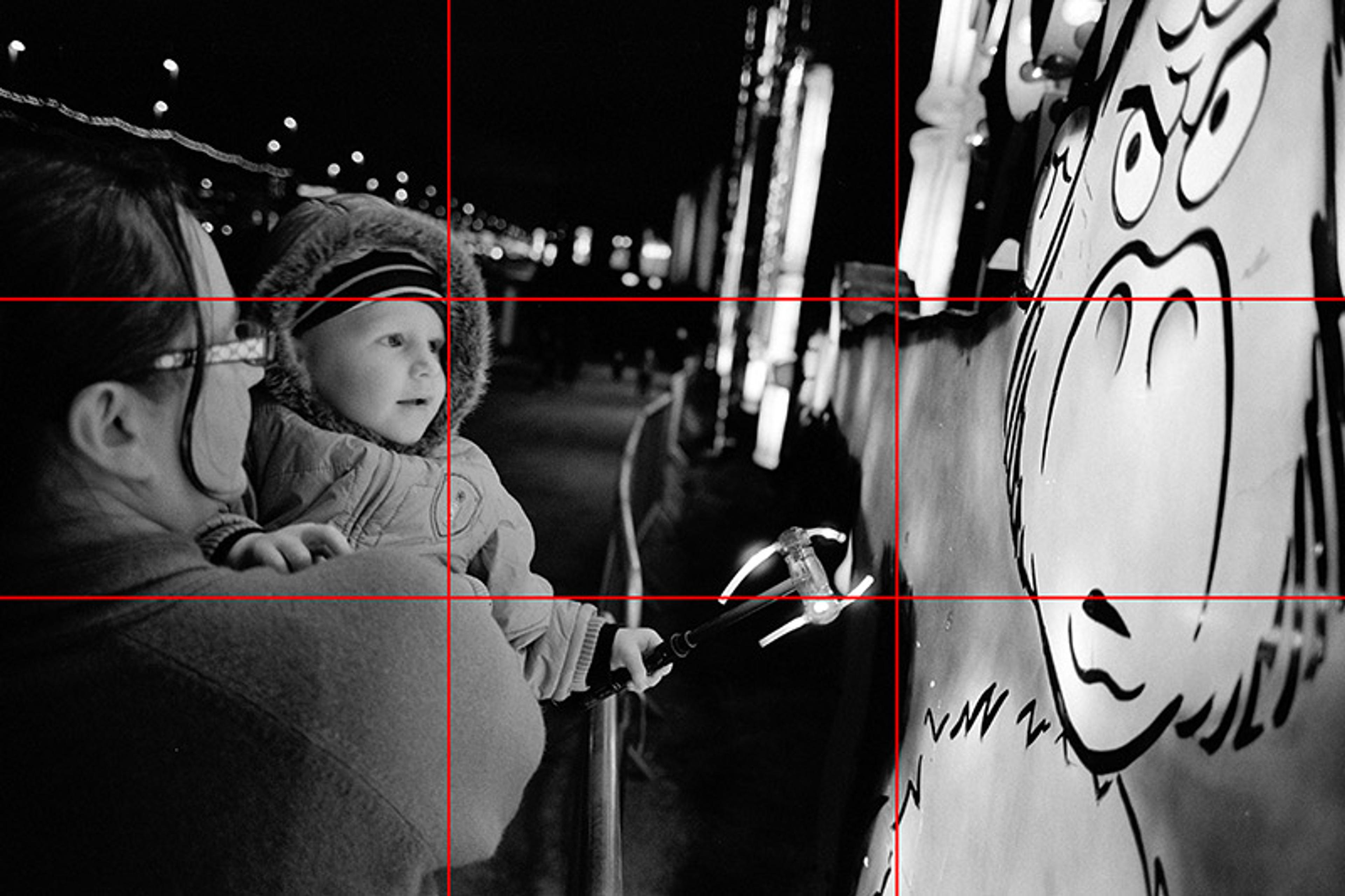 A woman holds a young child who is looking at a cartoon figure on a poster. The image is overlayed with a grid of 9 rectangles dividing the picture into sections