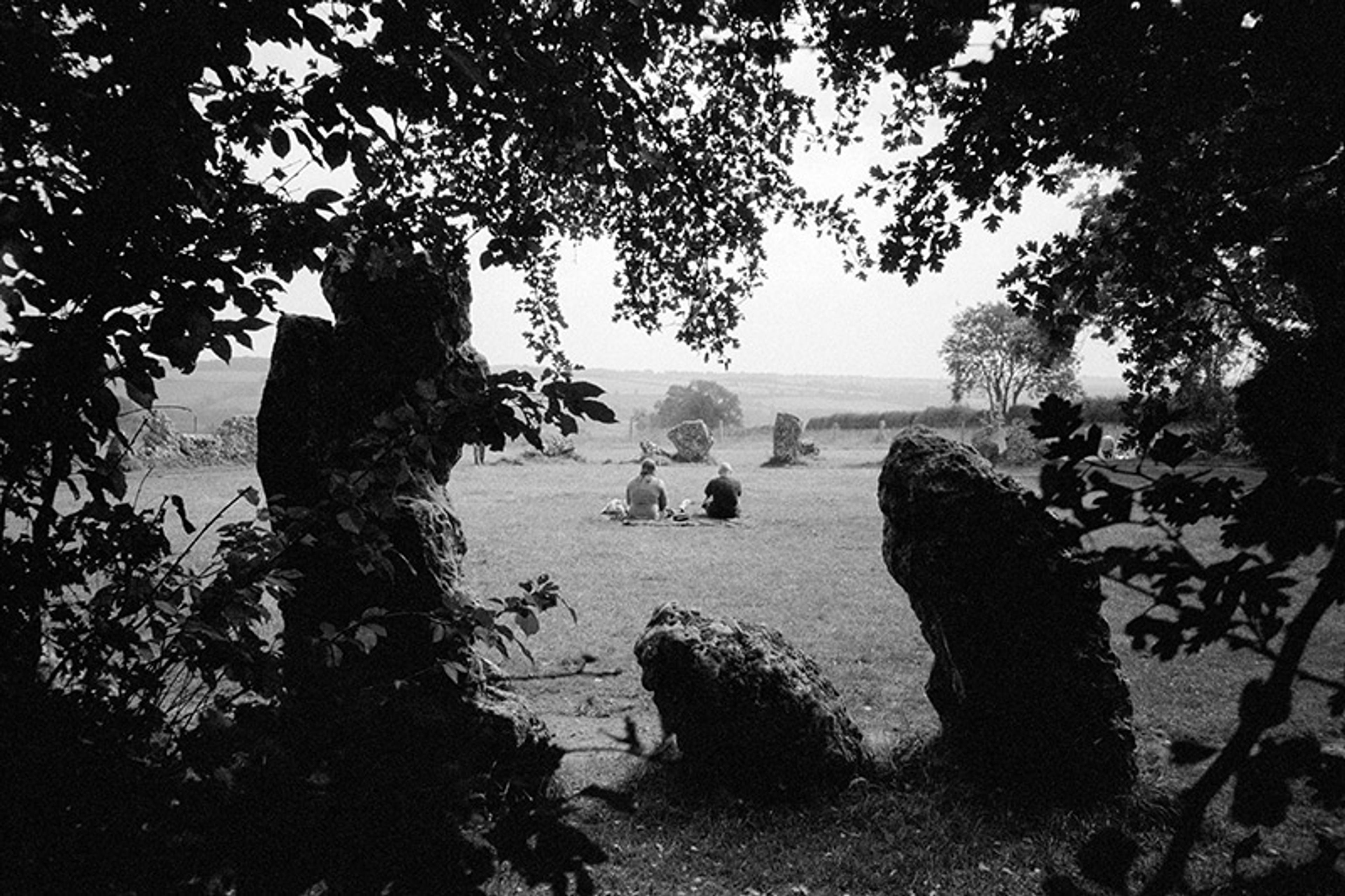 A black-and-white image of a couple seated in the centre of an ancient stone circle. The image is shot through dark foliage such that the foreground frames the couple