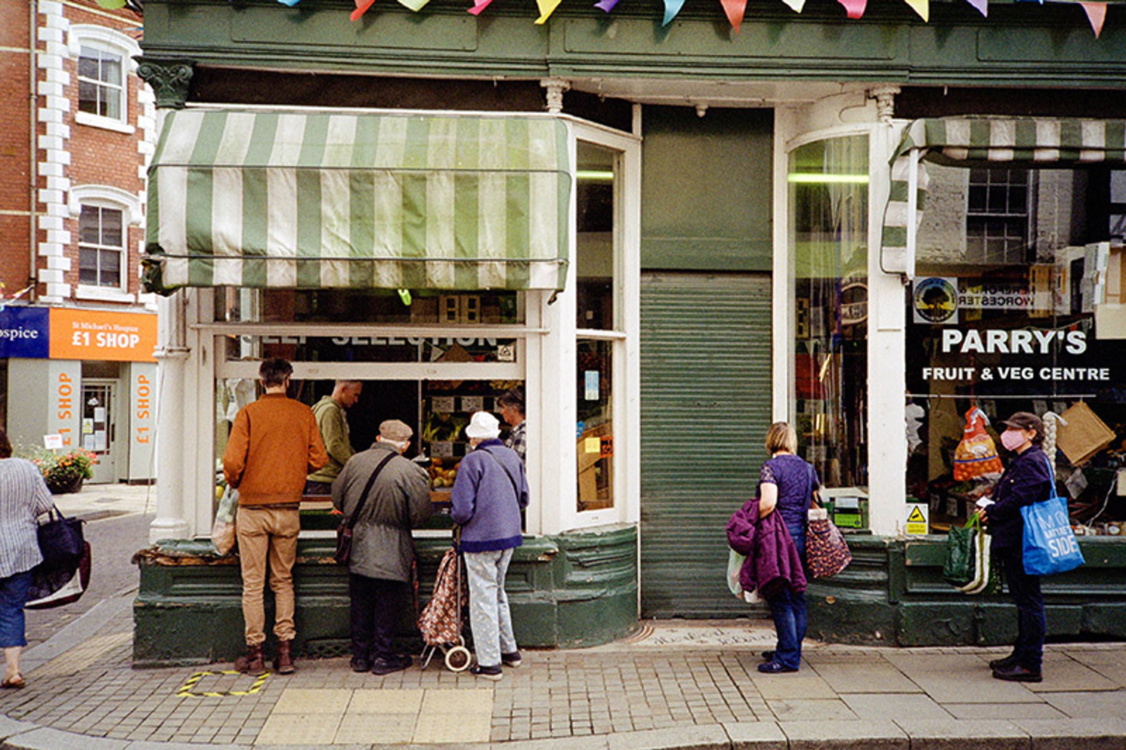 People of differing heights and ages queue outside a traditional greengrocer’s shop