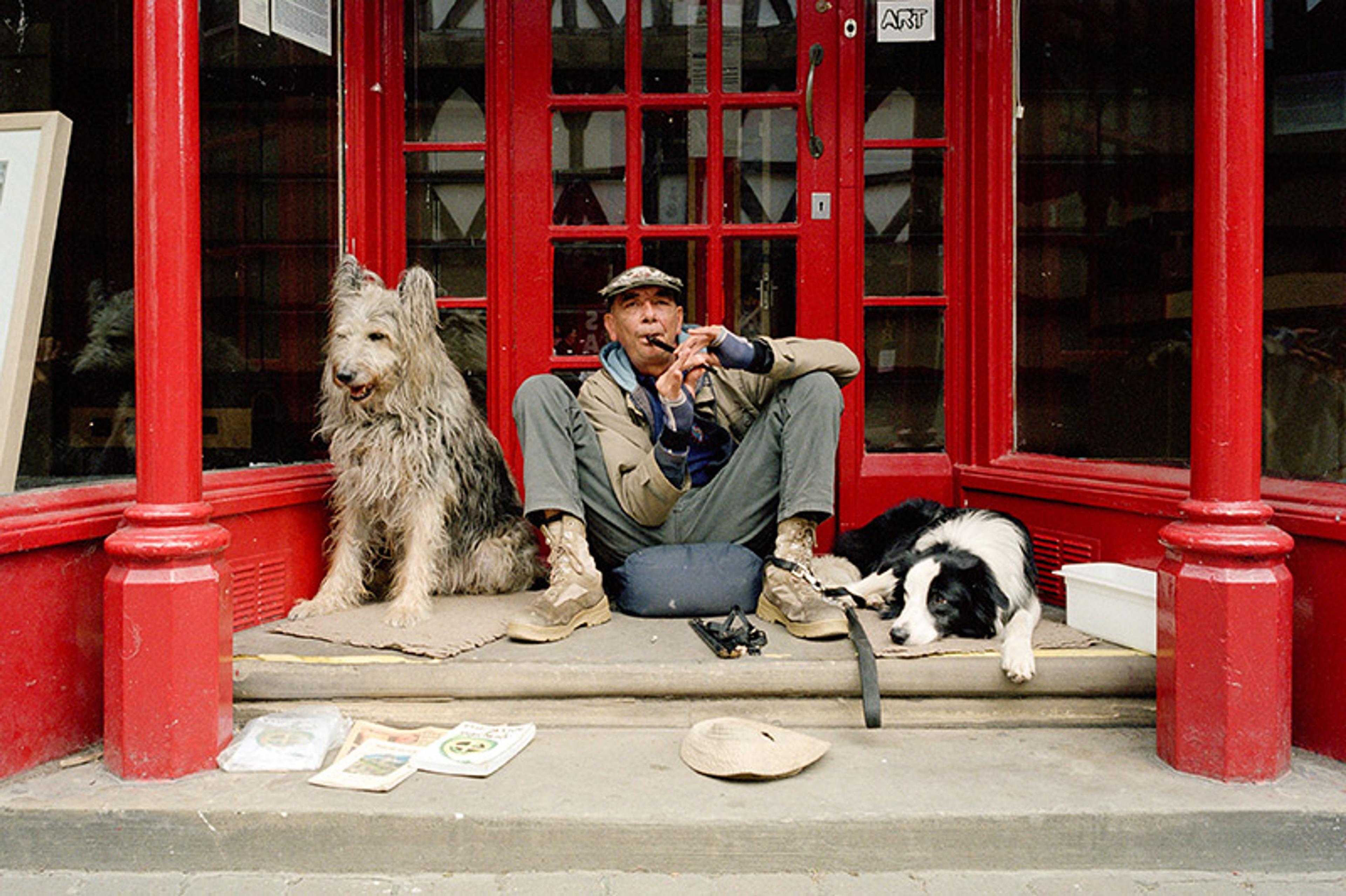 A busker sits in a red-painted shop doorway playing a tin whistle. Alongside him are his two dogs