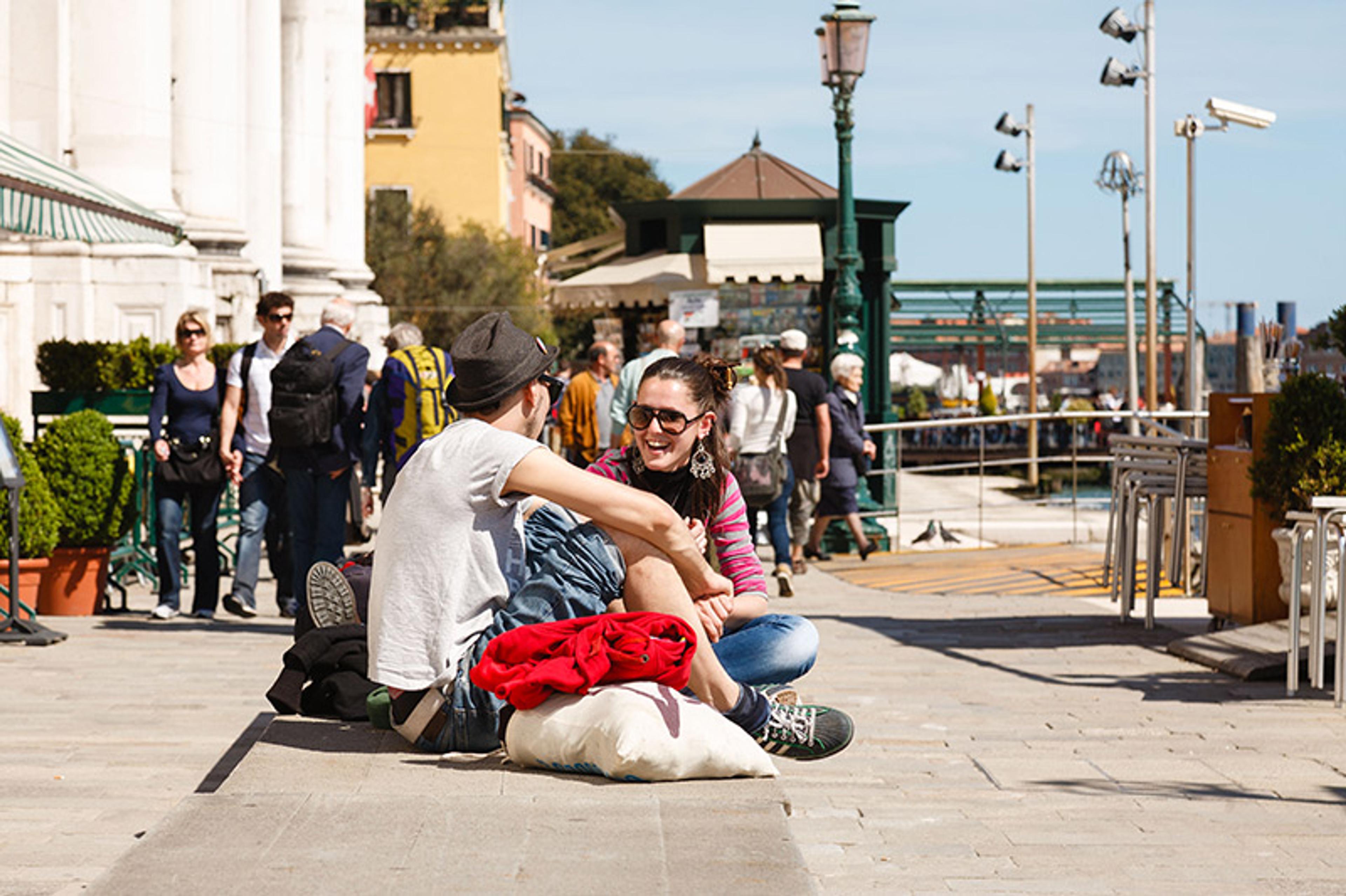 A happy young couple sitting and chatting on a wall in the sunshine. They are in focus while people in the background are defocused