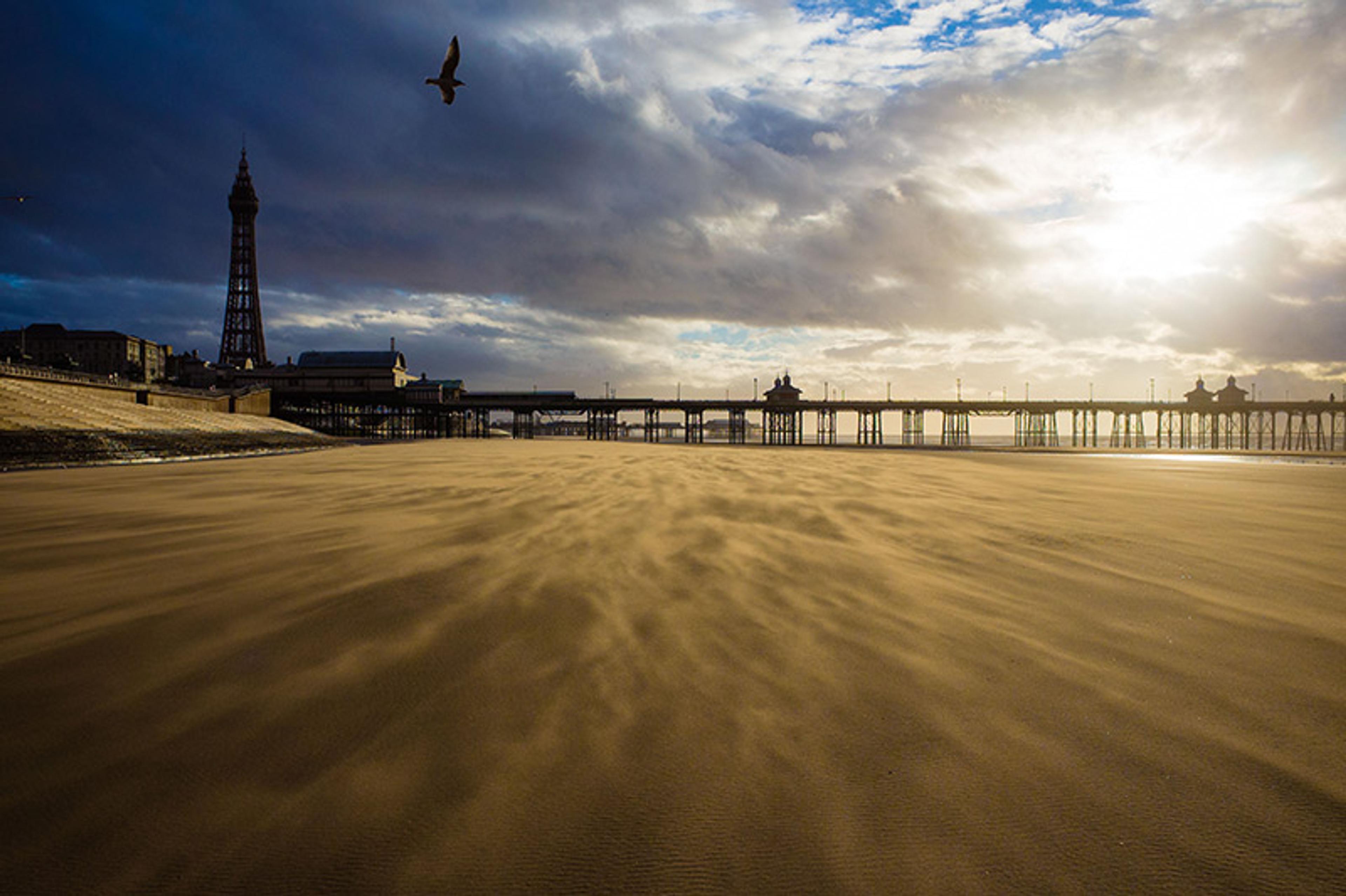 A side view of Blackpool pier seen from the beach at low tide. The sun is bursting through late afternoon clouds and the wind is forming patterns in the sand