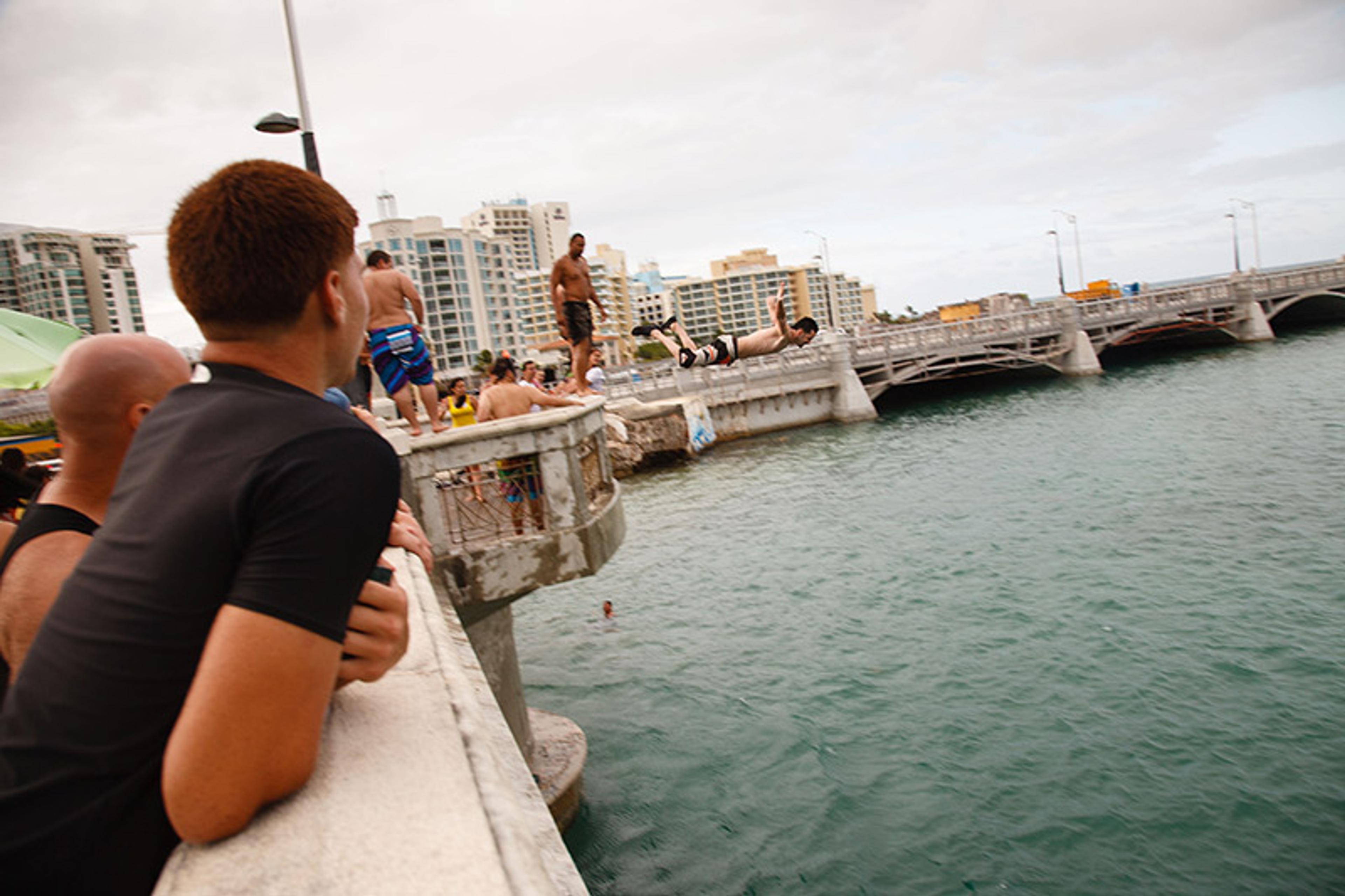Spectators watch people diving off an ancient bridge into a harbour. In the background are modern apartment buildings