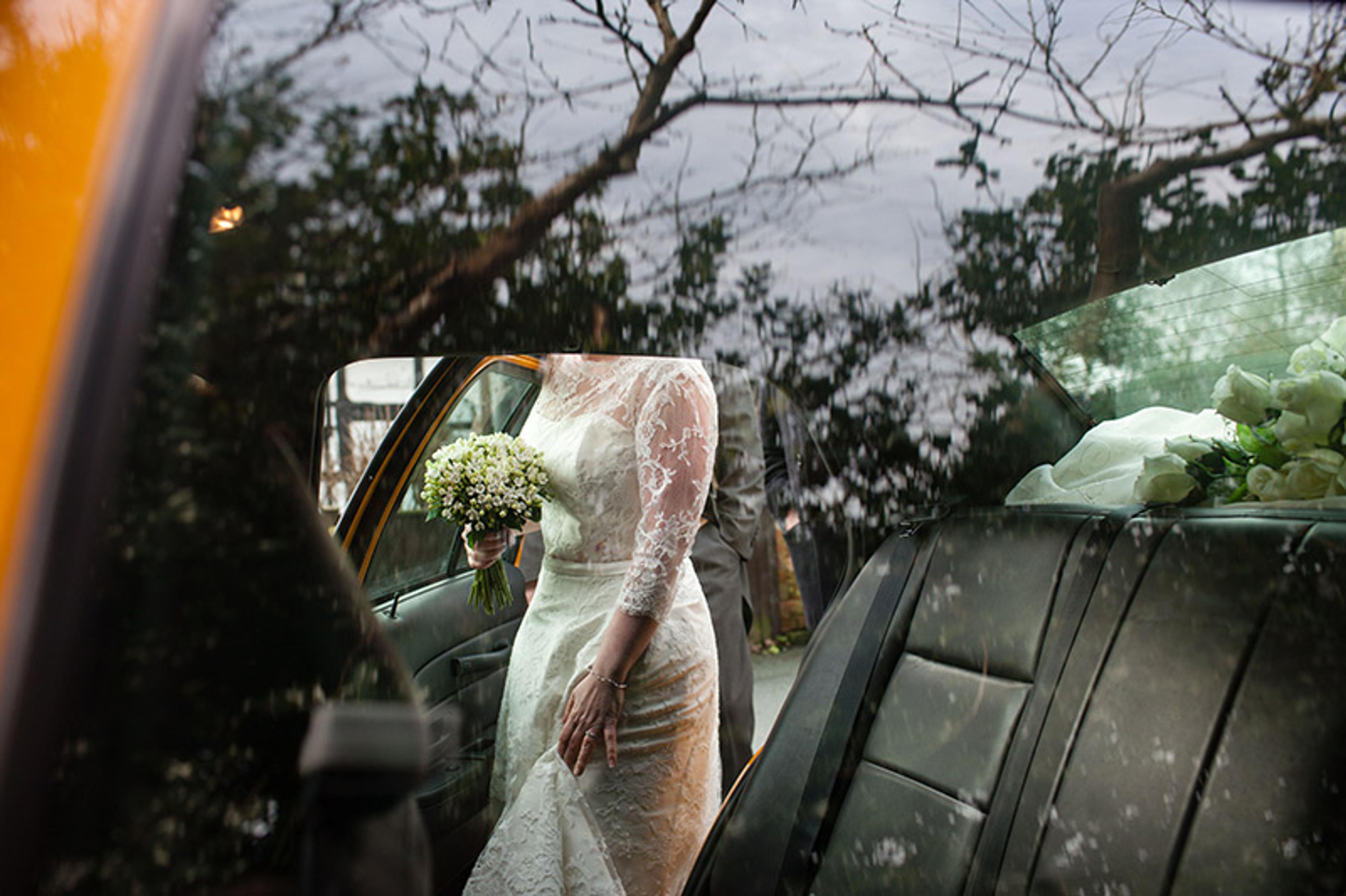 A bride holding flowers is seen about to get into a car. The photo is taken through the glass of the car window on the opposite side of the car and the bride’s head is not visible. There are numerous reflections in the glass