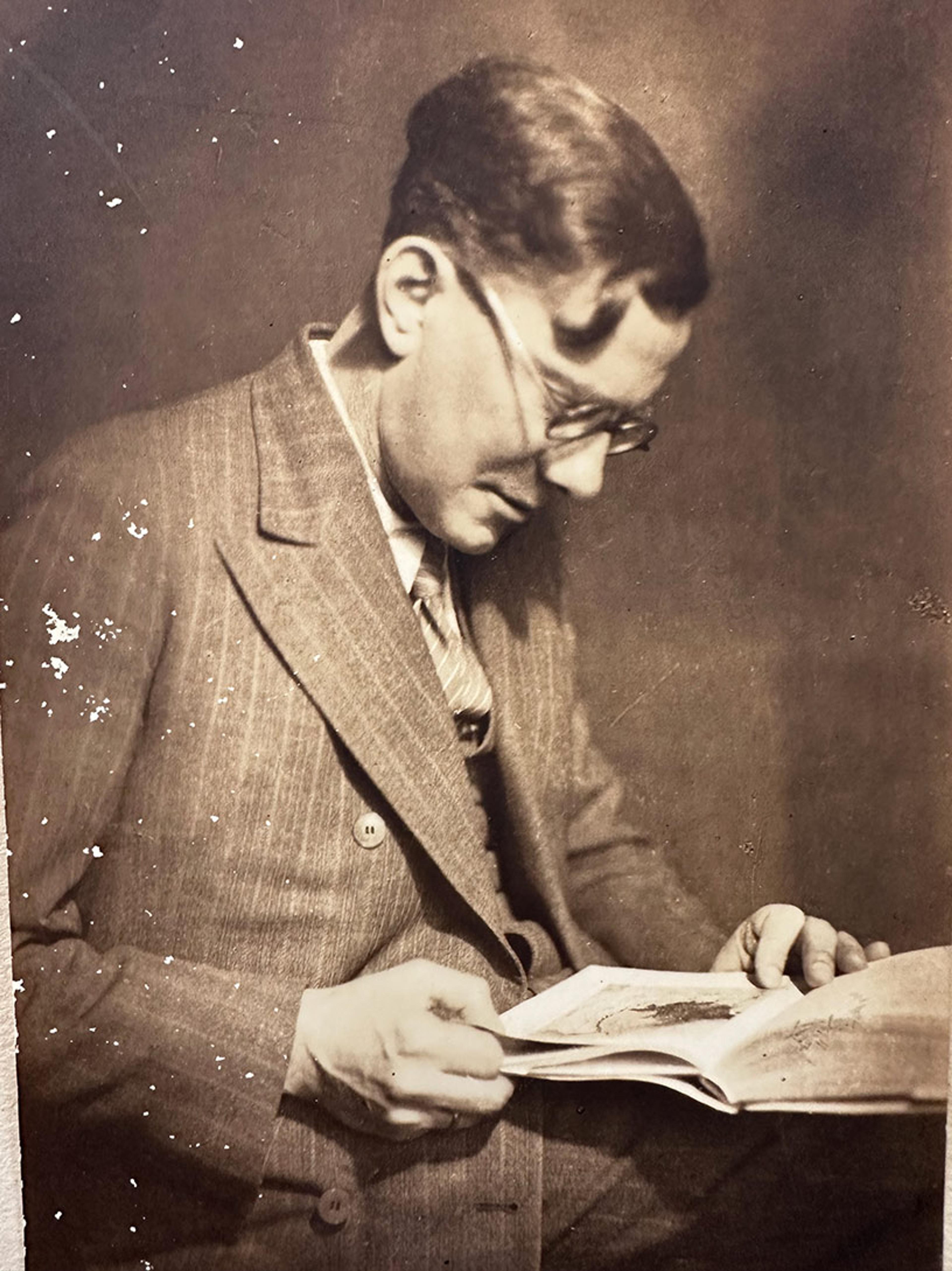 A sepia-tone photograph of a man with glasses and wavy hair, dressed in a pinstriped suit with a tie, engrossed in reading an open book. The image has visible wear and speckling, giving it a vintage feel.